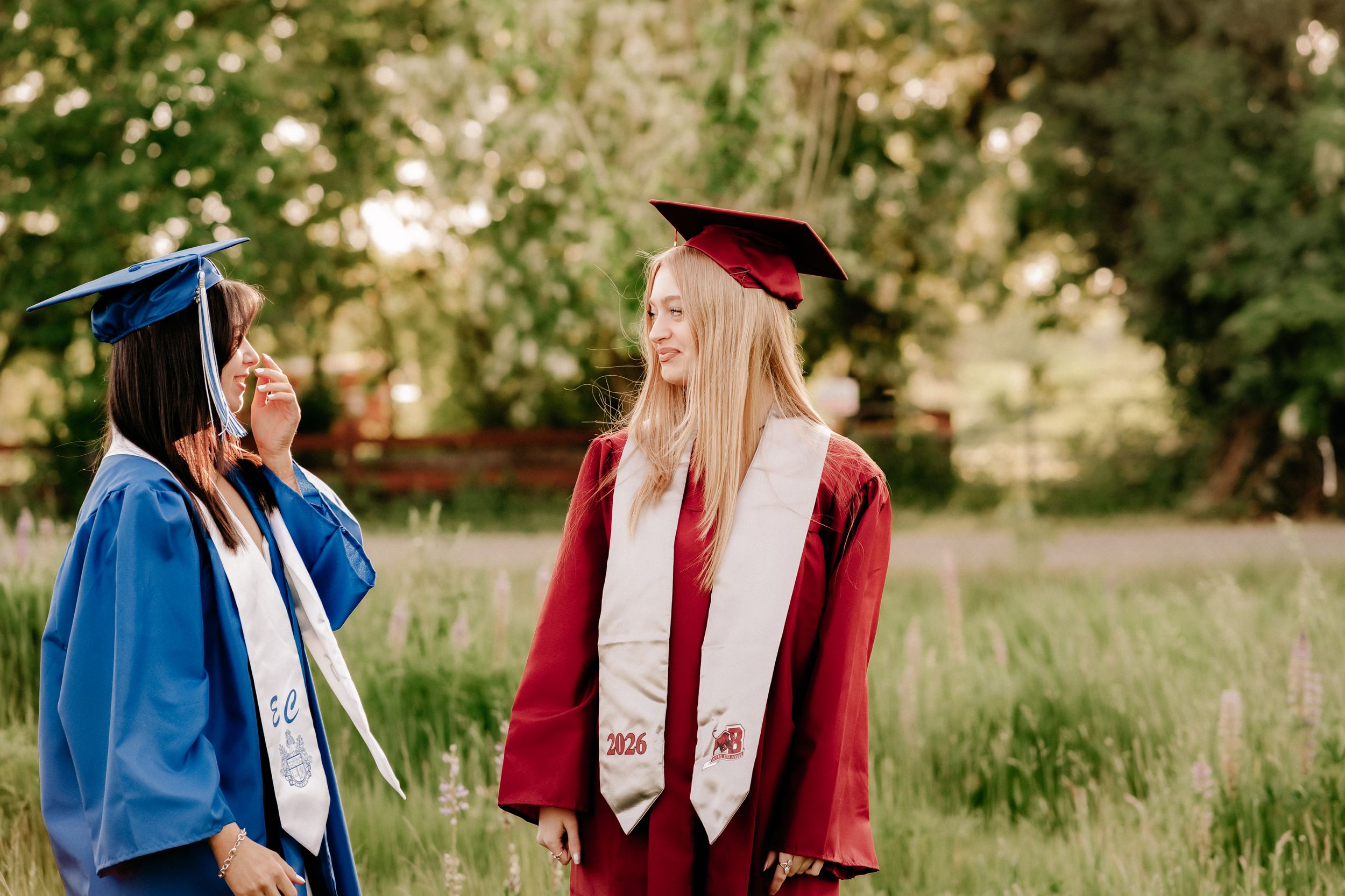 Two female graduates in blue and red cap and gown regalia smiling outdoors in a green field during senior photos in Puyallup, Wa.