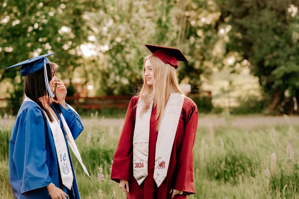 Two female graduates in blue and red cap and gown regalia smiling outdoors in a green field during senior photos in Puyallup, Wa.