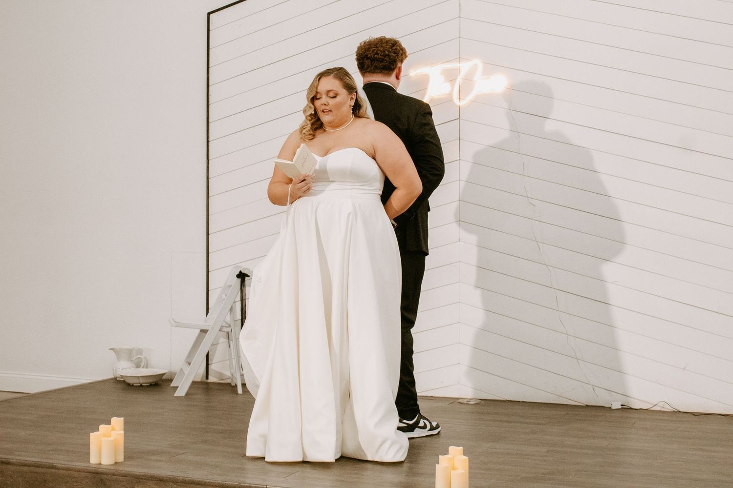 Bride in strapless white gown with groom in black suit at candlelit wedding ceremony with neon sign backdrop.