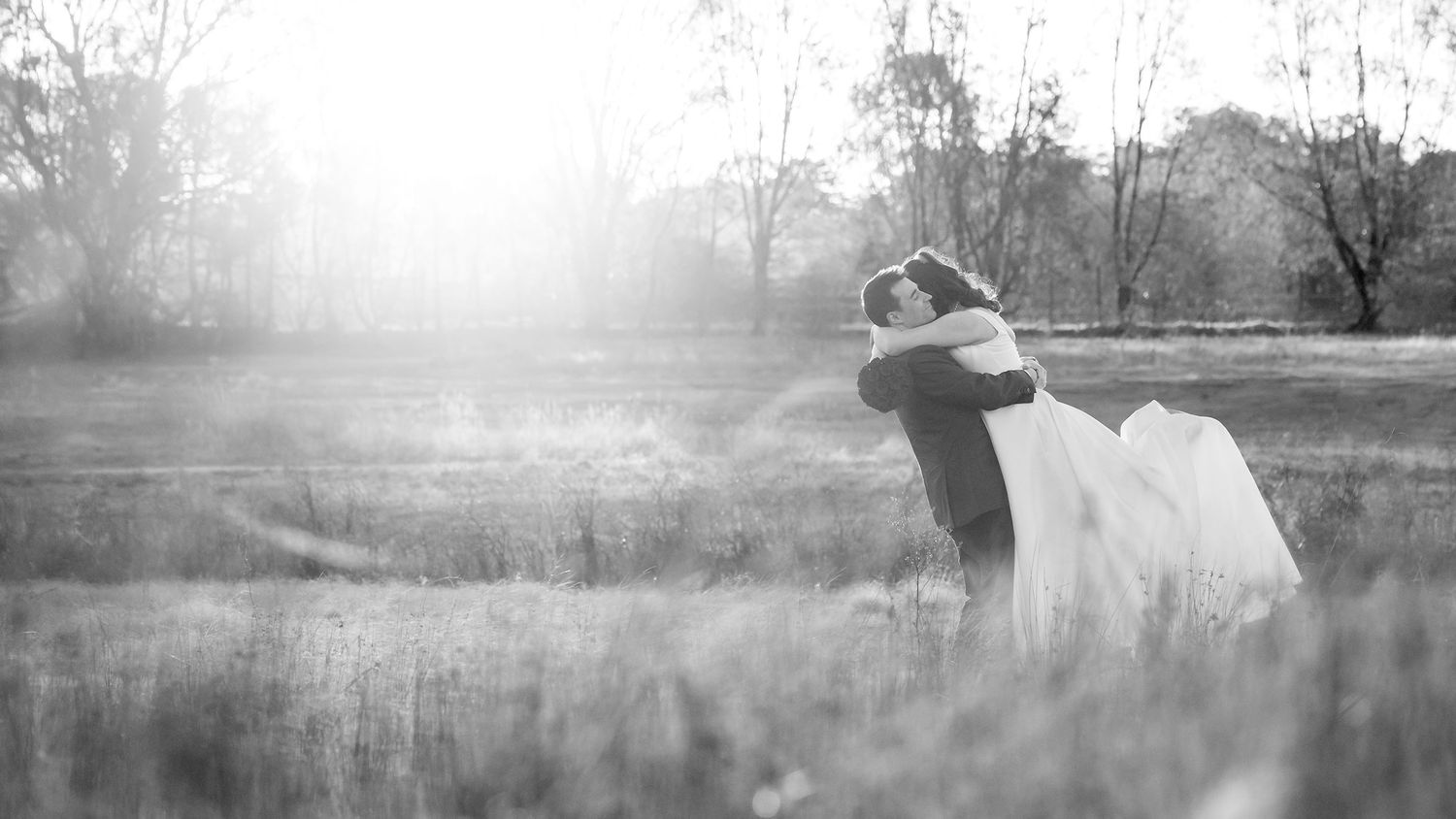 A black and white image of a Bride and Groom sharing a hug at Sunset in a field of long grass.