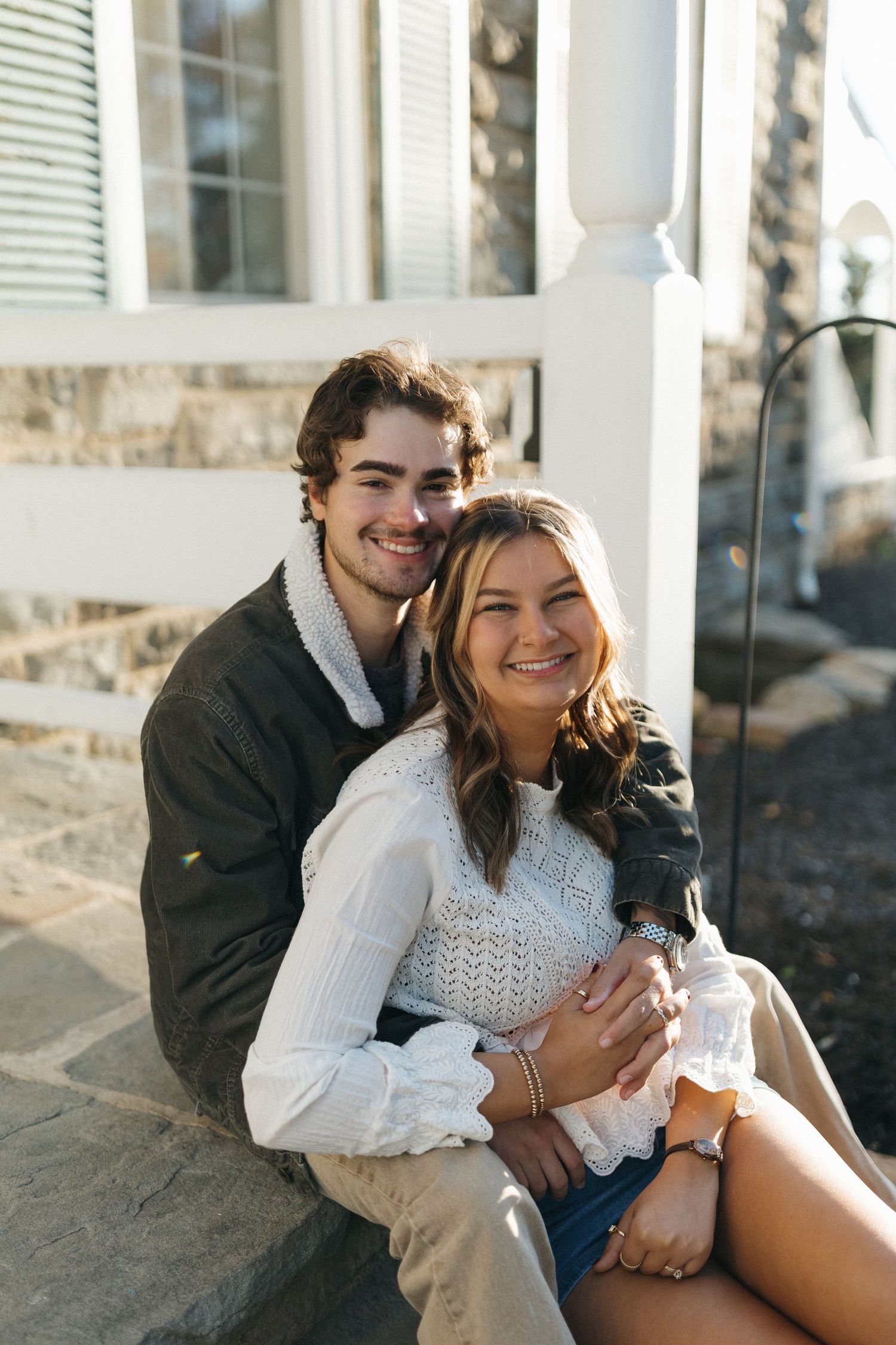 A couple sits together on stone steps outside a building, embracing and smiling on a sunny day.
