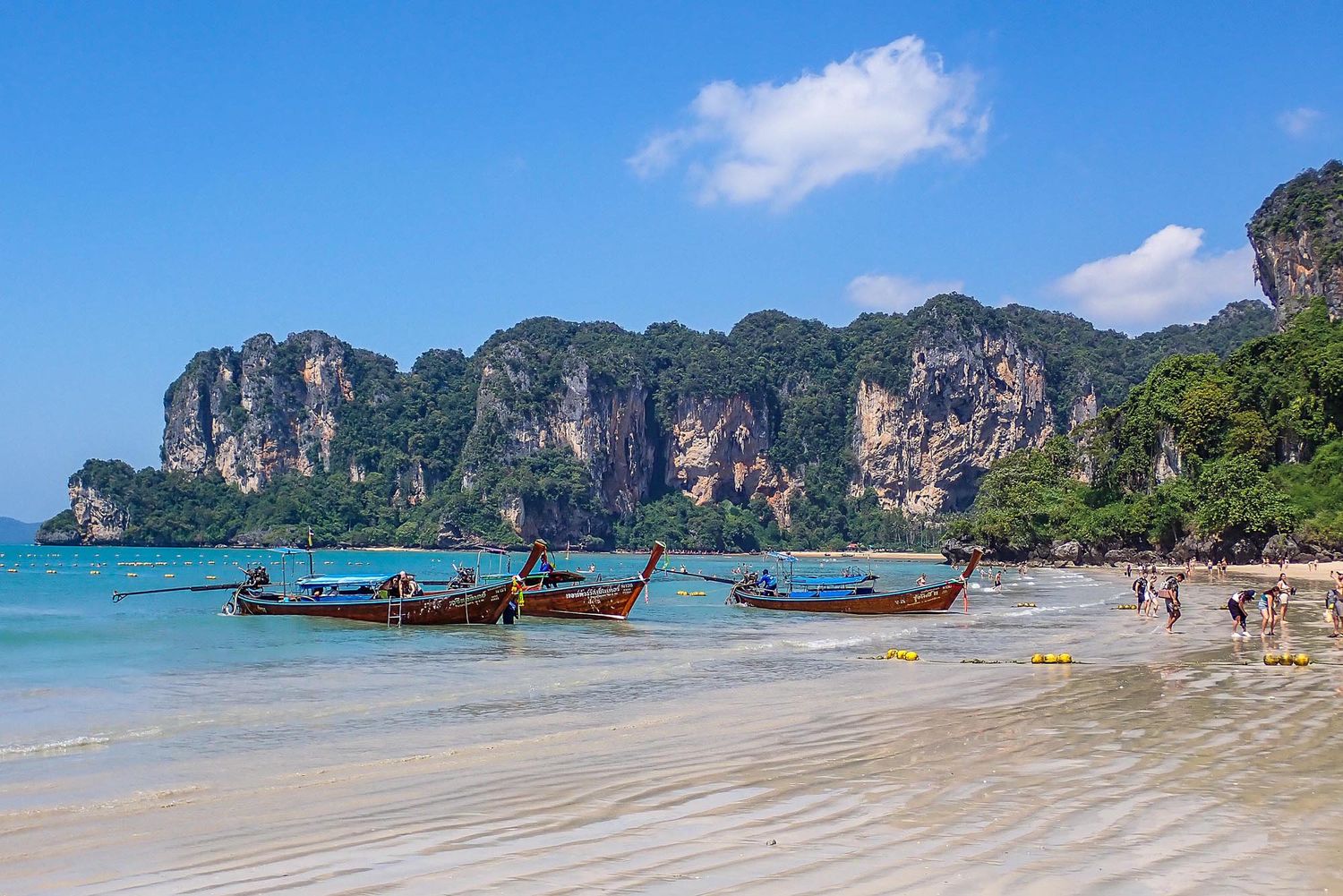 Traditional longtail boats float on turquoise waters near limestone cliffs at Railay Beach in Krabi, Thailand.