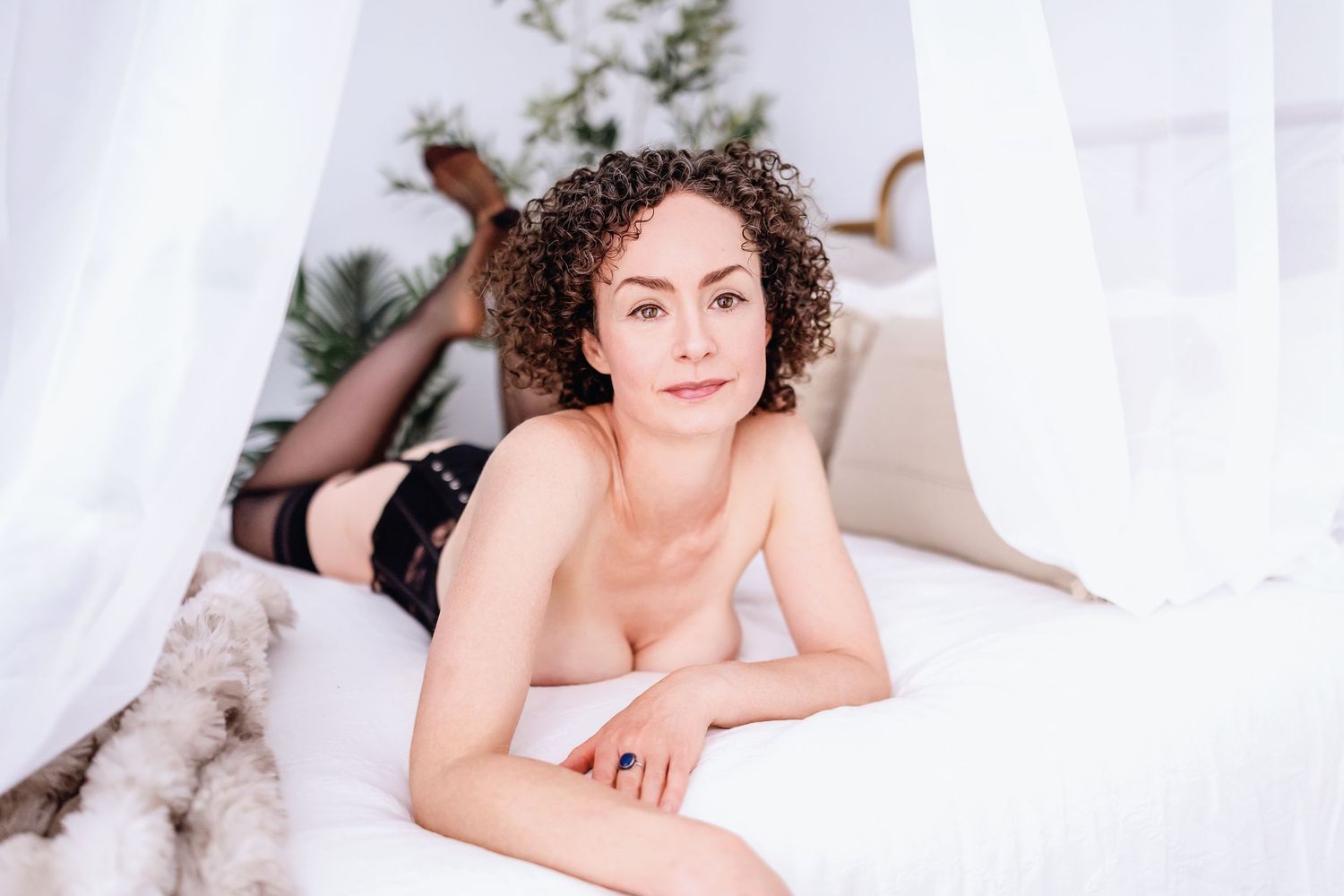 Person with curly hair lies gracefully on white bedding while looking at camera.