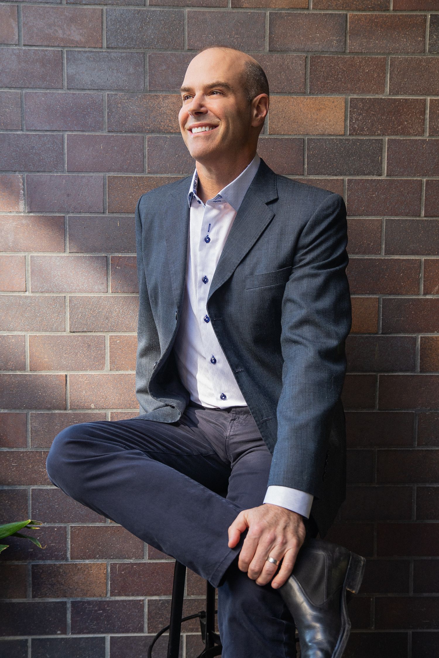 Professional headshot portrait of someone in a gray suit and light blue dress shirt against a brick wall background.