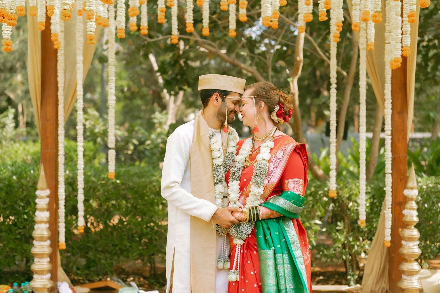 A couple in traditional wedding attire stands beneath a decorated mandap with hanging floral arrangements.