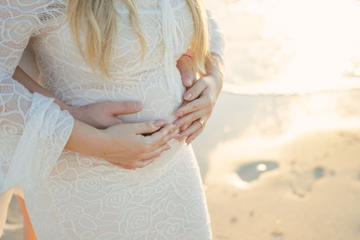 A touching maternity photo shows a person wearing a white dress cradling their baby bump against a golden sunset.