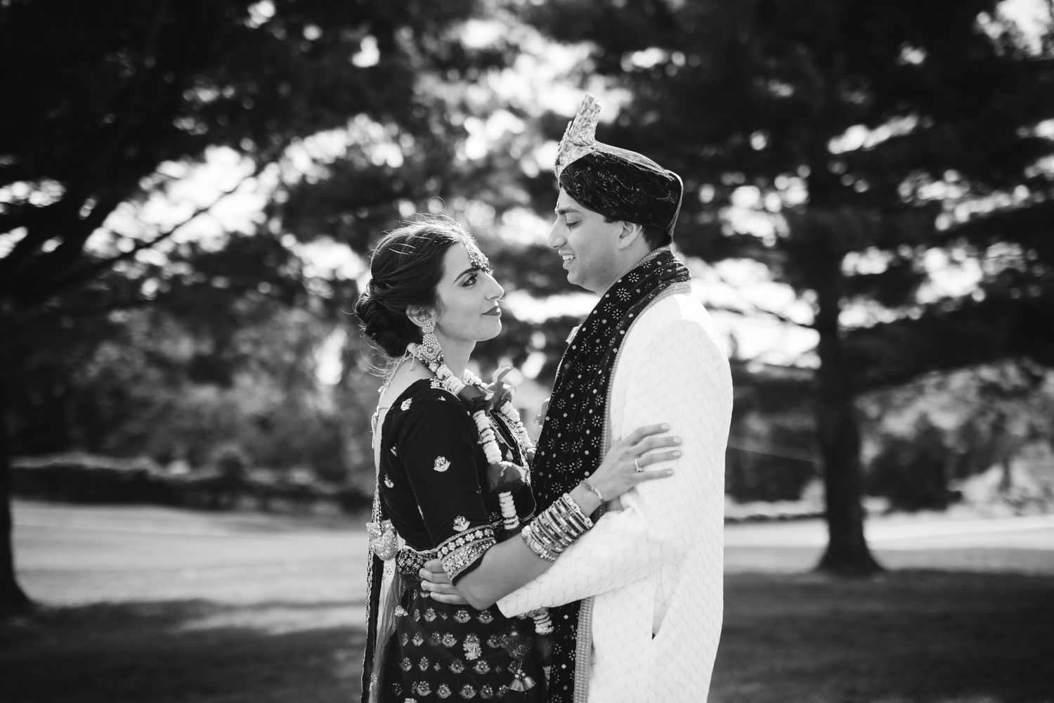 A romantic black and white portrait of a couple embracing outdoors in a park setting with trees in the background.