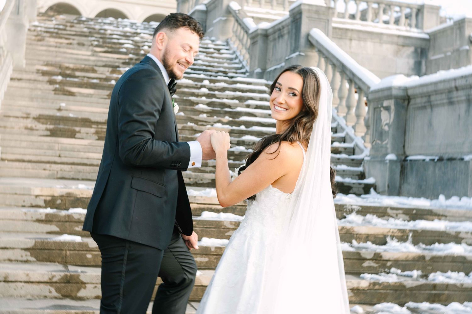 A hand wearing an engagement ring rests against a white lace wedding dress with delicate beaded straps.