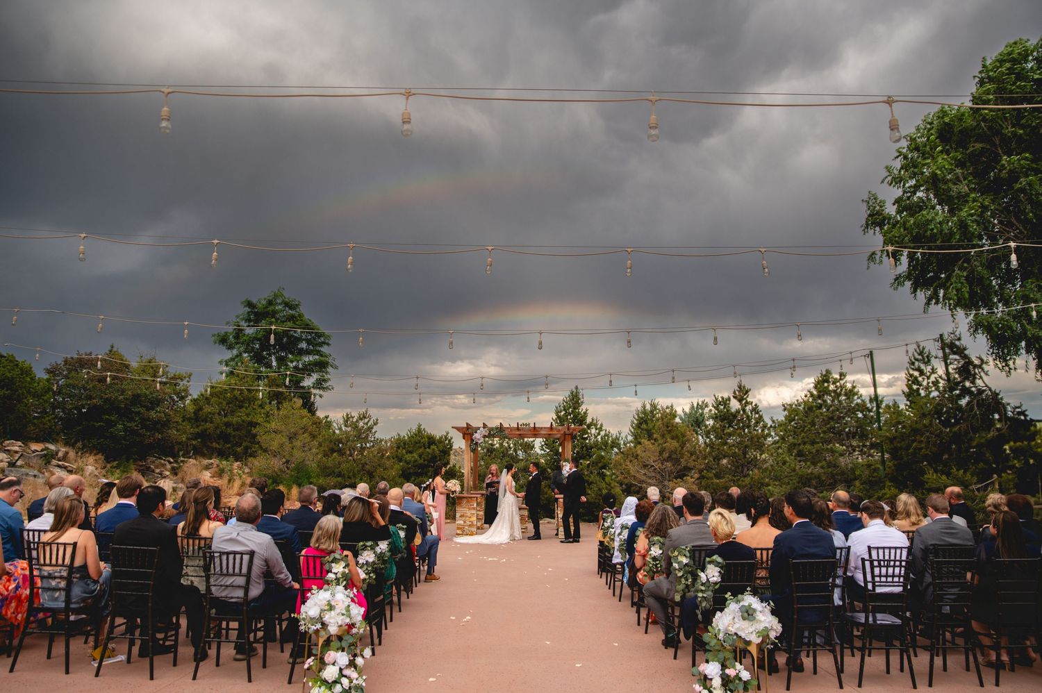 A scenic outdoor wedding ceremony setup with white chairs facing an altar under dramatic storm clouds at sunset.