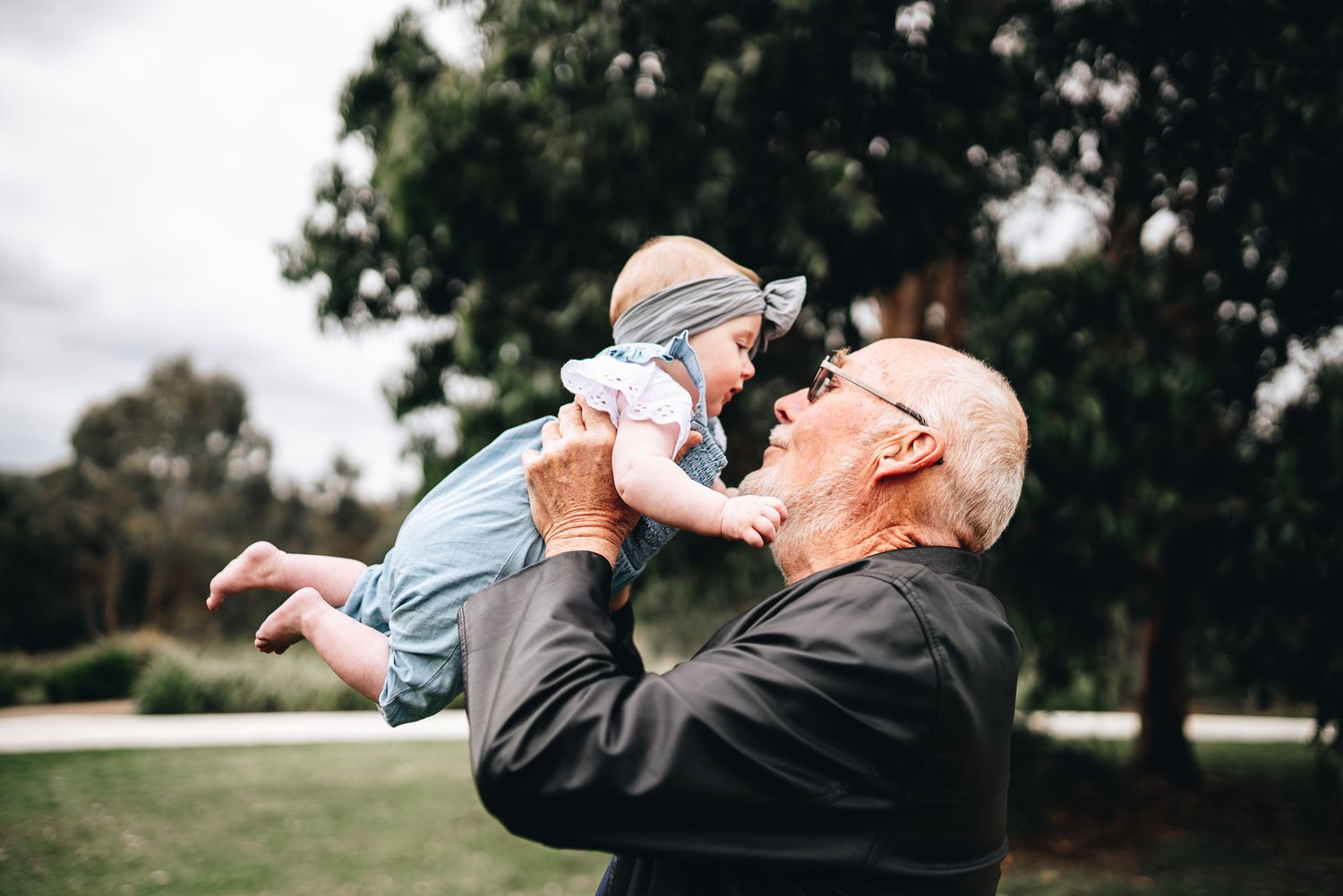 Grandfather in dark clothing holds up a small baby wearing light blue in an outdoor park setting taken by Melbourne family photographer.