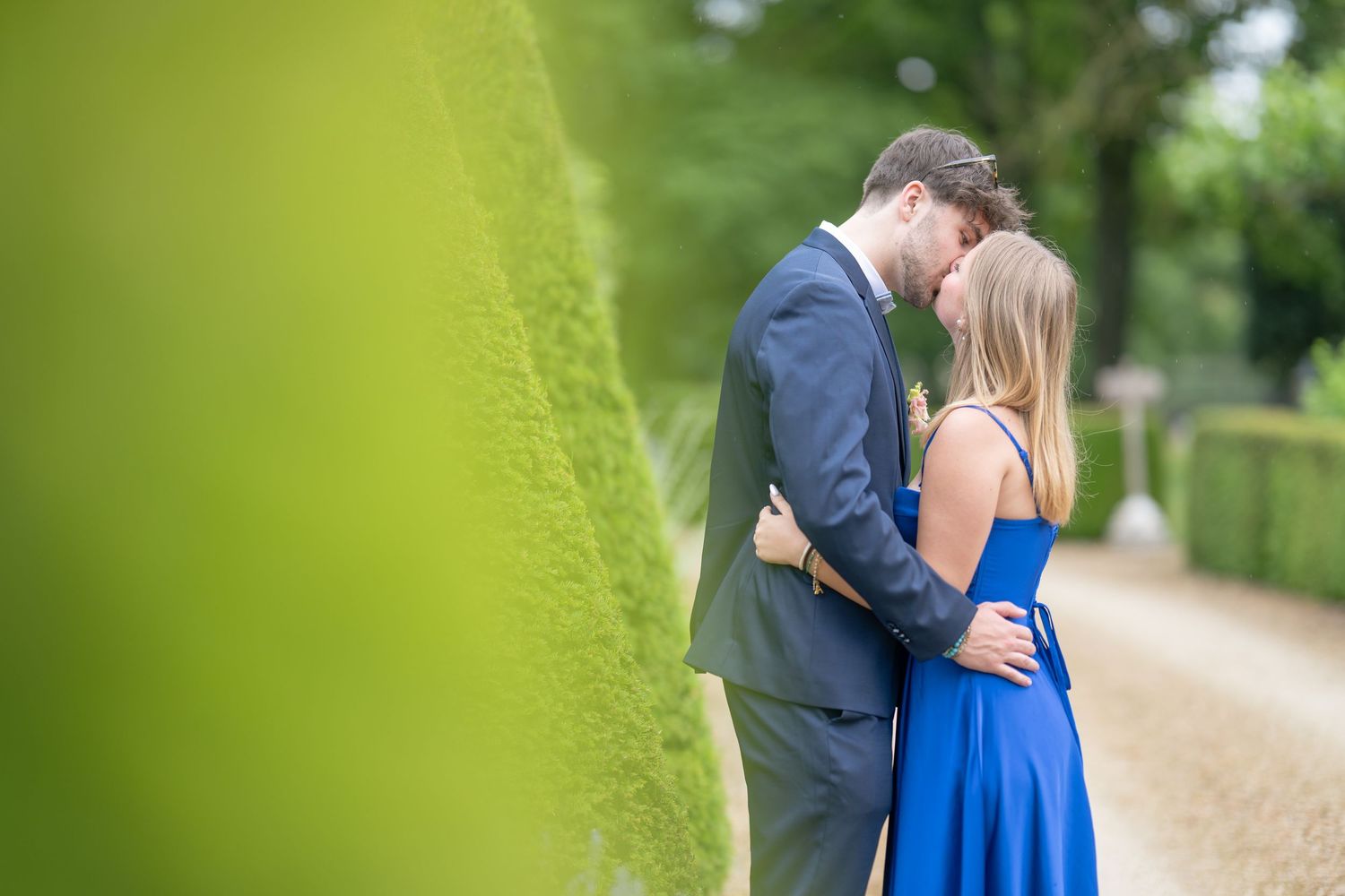 A romantic couple embraces on a garden path with green hedges creating a soft blurred frame around them.