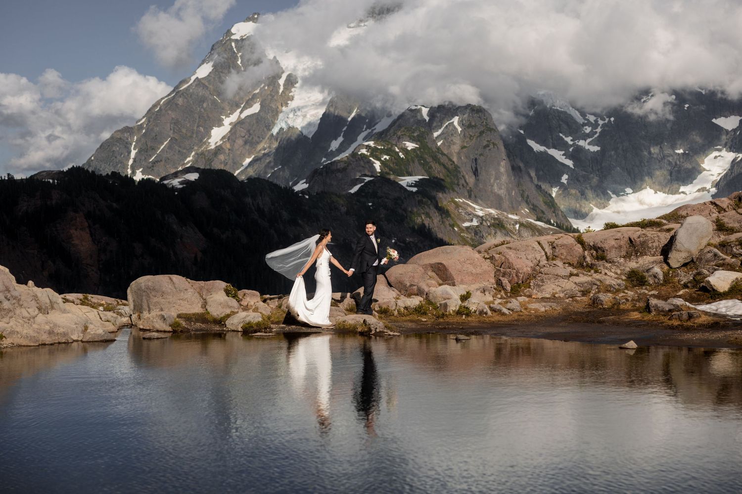 A couple in wedding attire poses against a dramatic mountain backdrop with snow-capped peaks and a serene alpine lake.