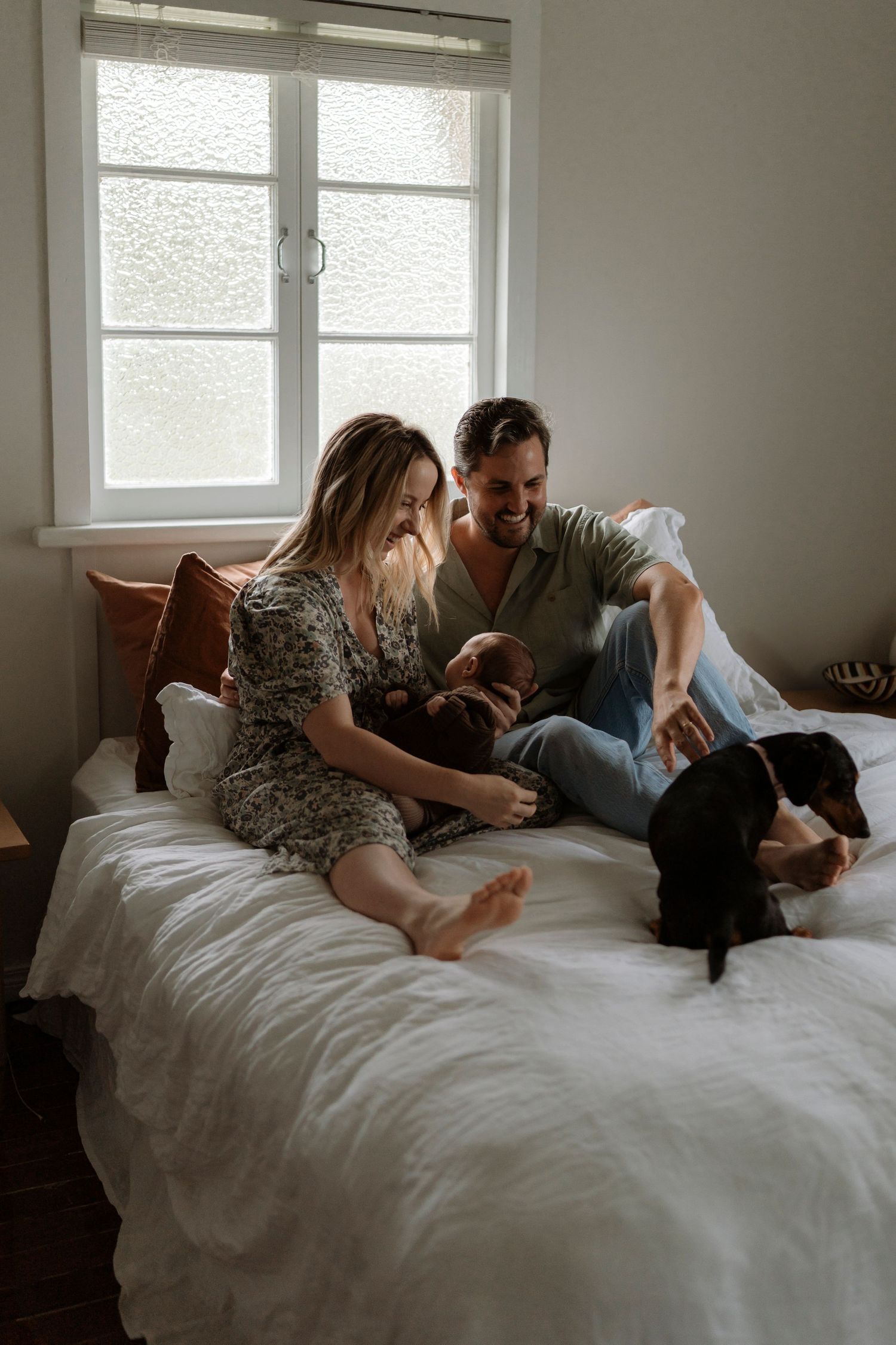 Three cozy photos show a newborn family relaxing on a white bedspread in a sunlit room with windows in the background.