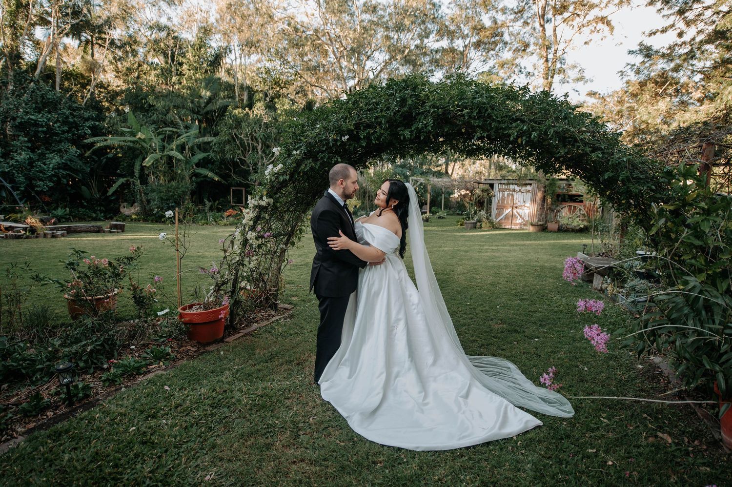 A couple shares an intimate moment under a lush garden archway decorated with greenery at an outdoor wedding ceremony.