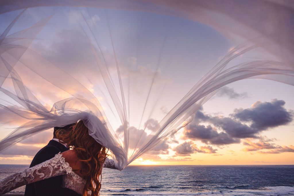 A flowing white fabric billows against a dramatic ocean sunset with clouds and sunbeams over the water.