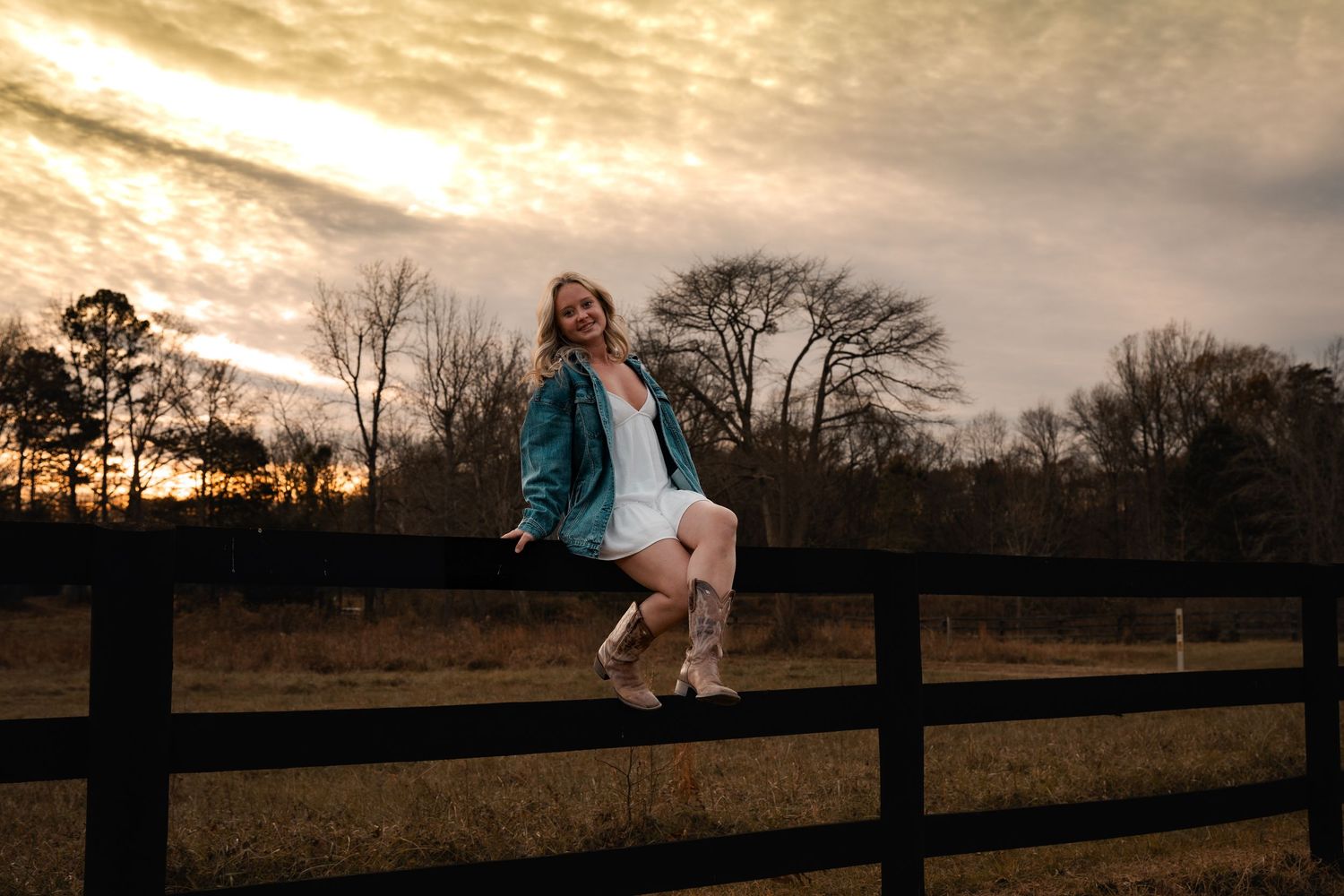 A person in a teal jacket sits on a wooden fence at sunset with silhouetted trees in the background.