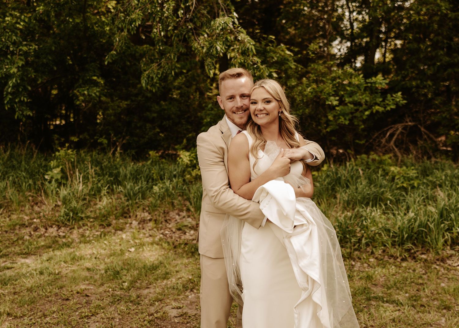 A newlywed couple embraces outdoors in a natural garden setting with warm sepia-toned lighting.