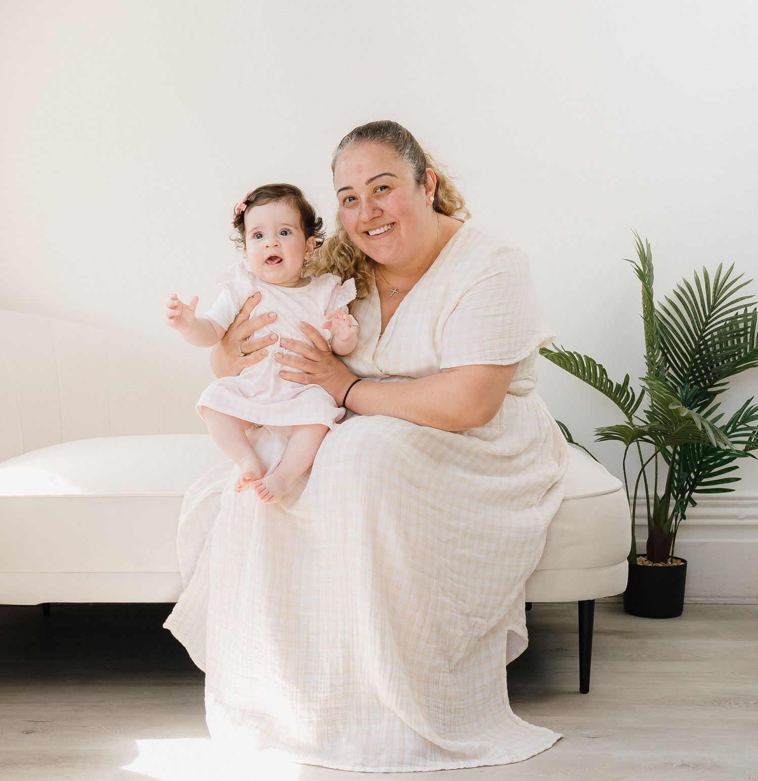 Mother and baby in white dresses sit together on floor in bright minimalist room with indoor plant.