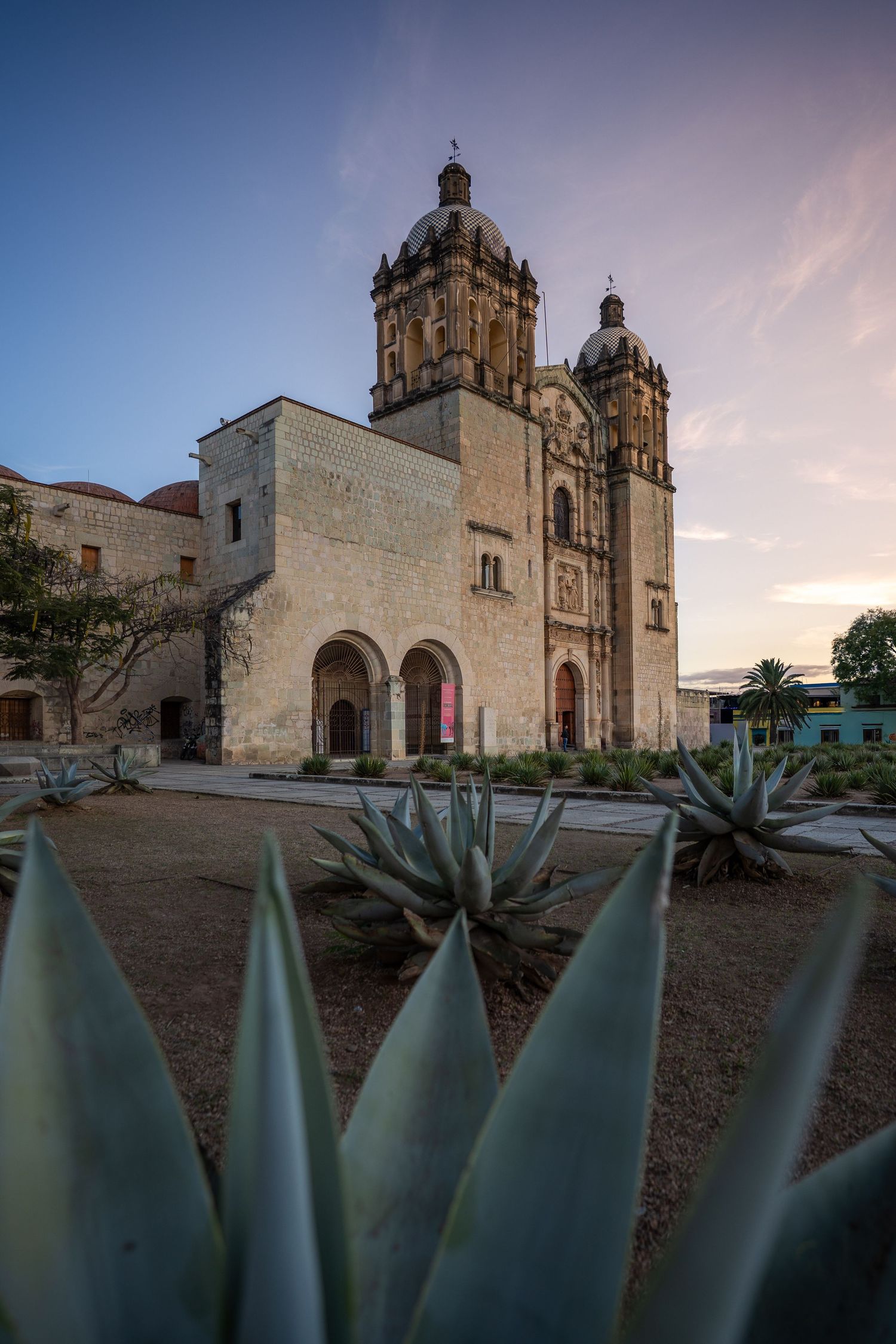Historic Spanish colonial church with agave plants in foreground against dramatic sky at sunset in Oaxaca, Mexico.