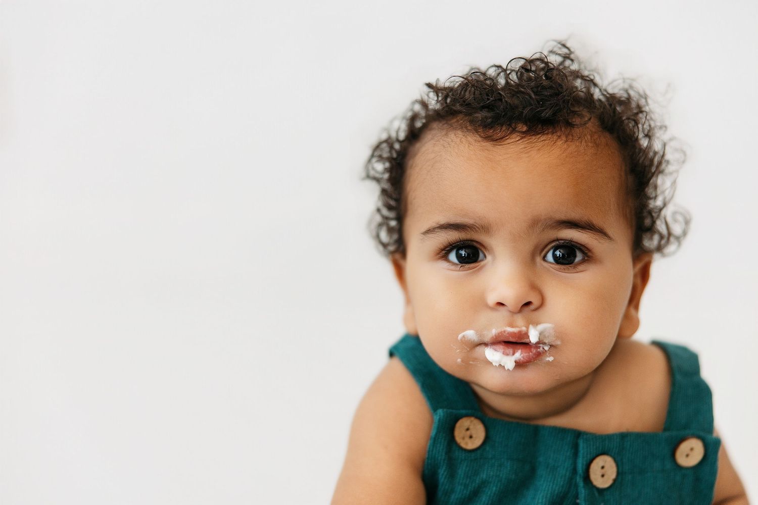 Small child wearing a green overall with messy white frosting or milk around their mouth against white background.