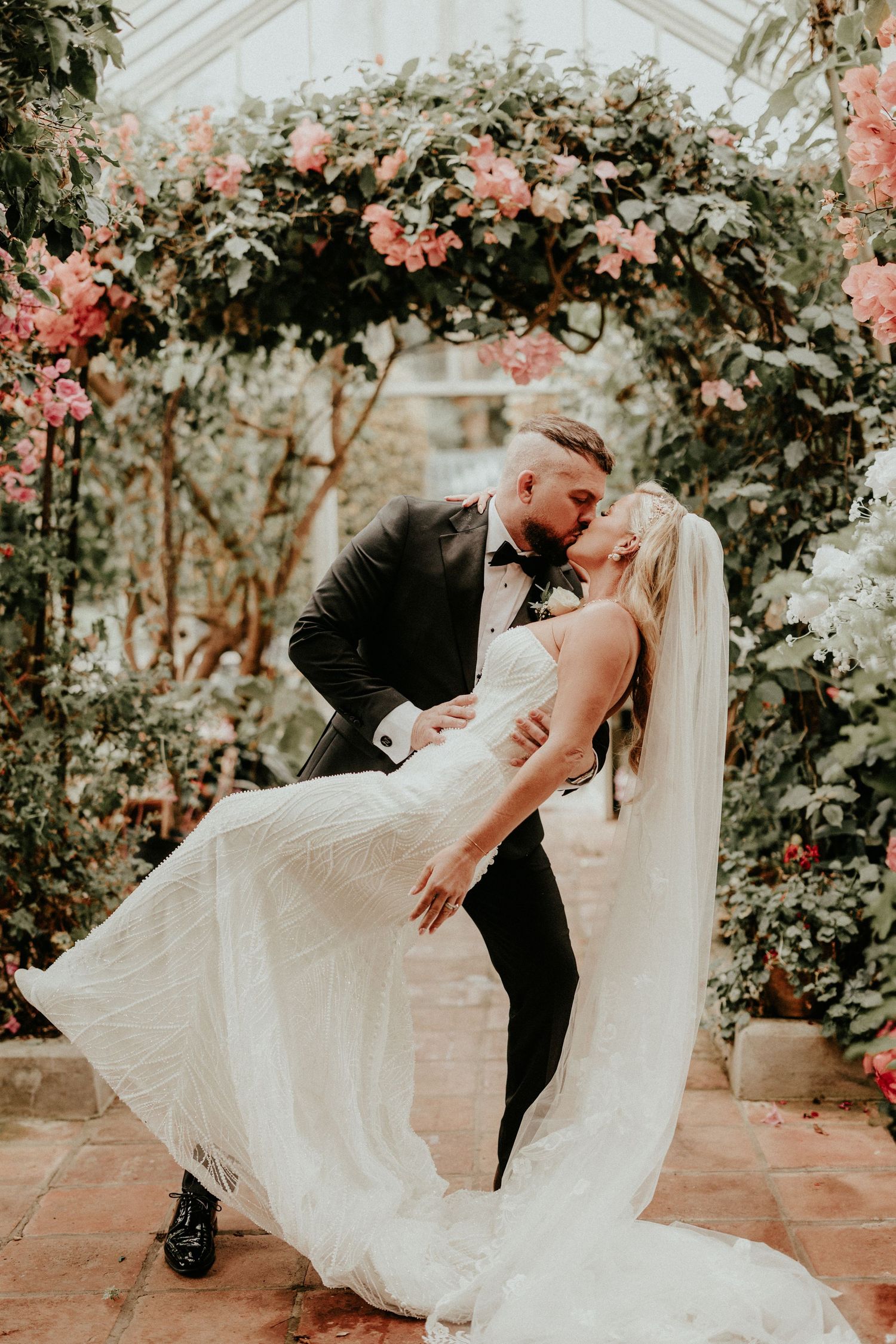 A romantic wedding couple dances together in a greenhouse-like setting surrounded by blooming flowers and natural light.