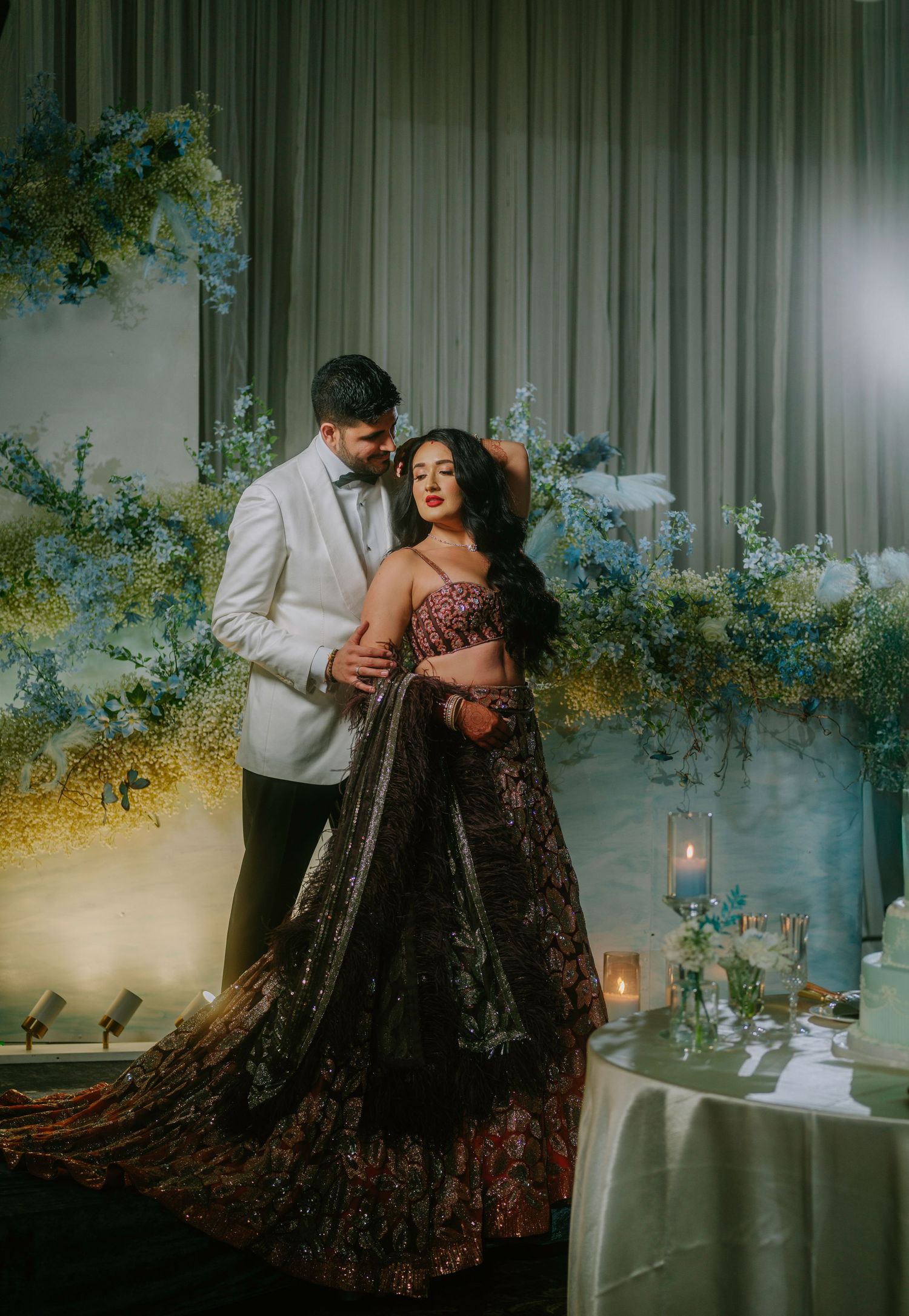 Romantic couple in elegant wedding attire pose intimately against a backdrop of blue floral decor and candlelight.