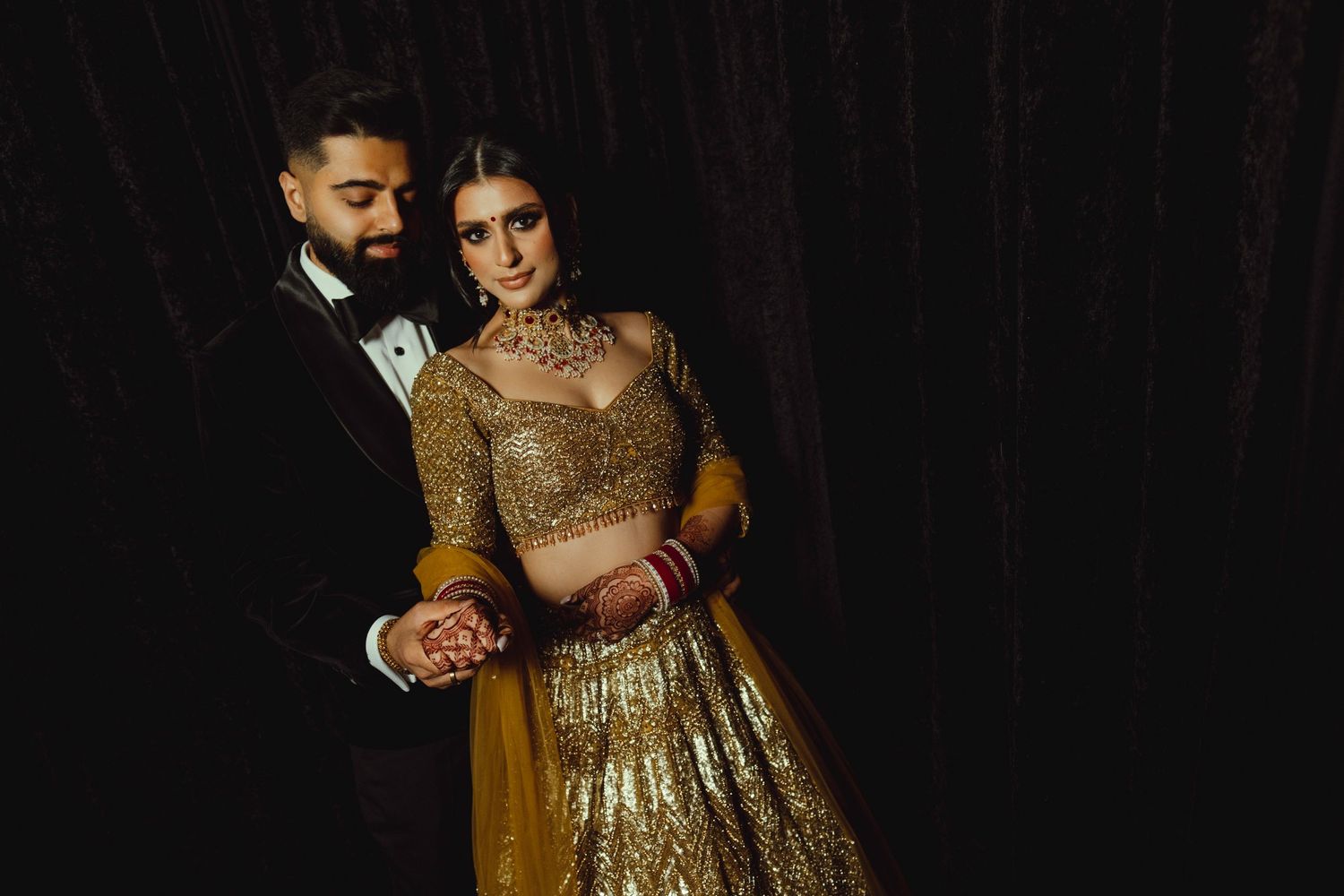 A couple in formal attire embrace in a dark setting, with the woman wearing a stunning gold sequined gown.