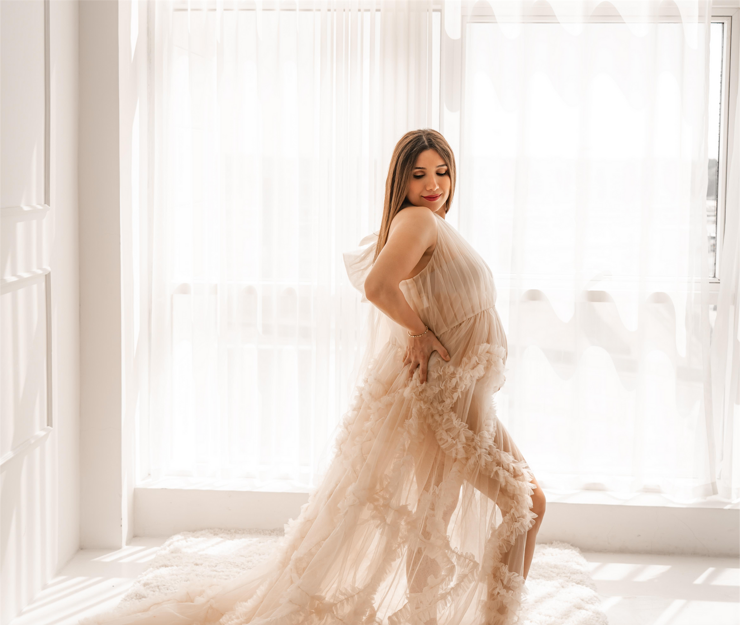 Maternity portrait in flowing sheer dress by bright window.