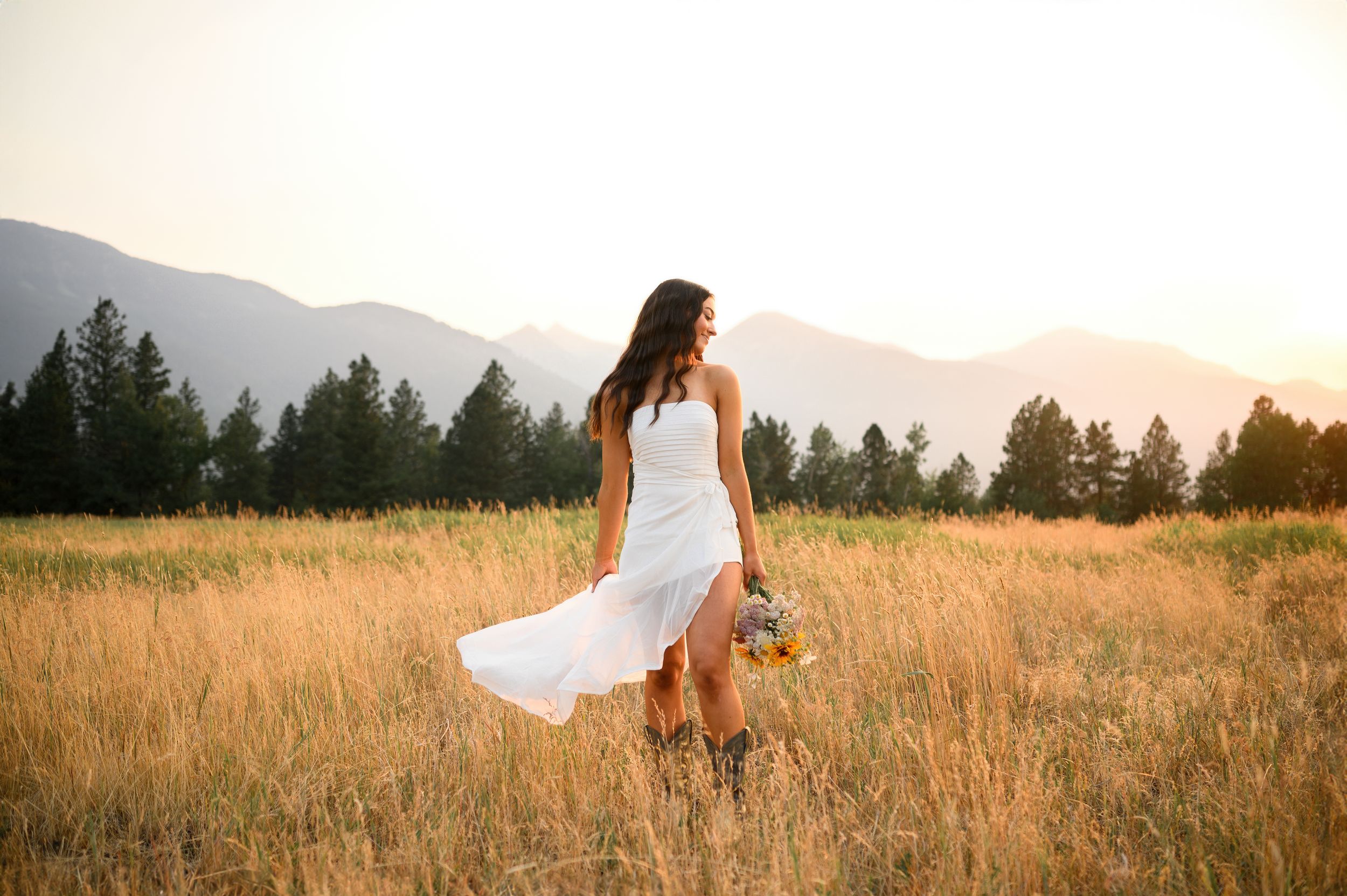 Woman in flowing white dress walks through golden field at sunset with mountain landscape in background.