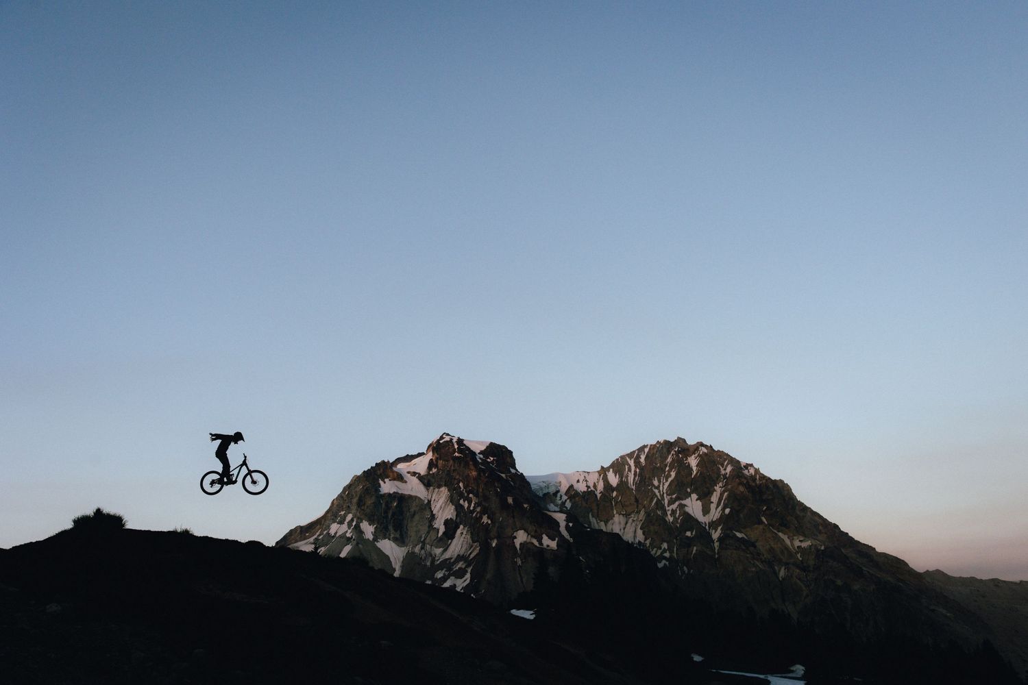 A silhouetted mountain biker jumps against a dramatic mountain landscape at dusk.