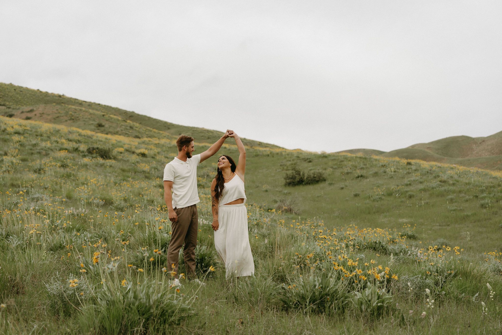 A couple shares a romantic moment in white attire while standing among wildflowers on a green hillside.