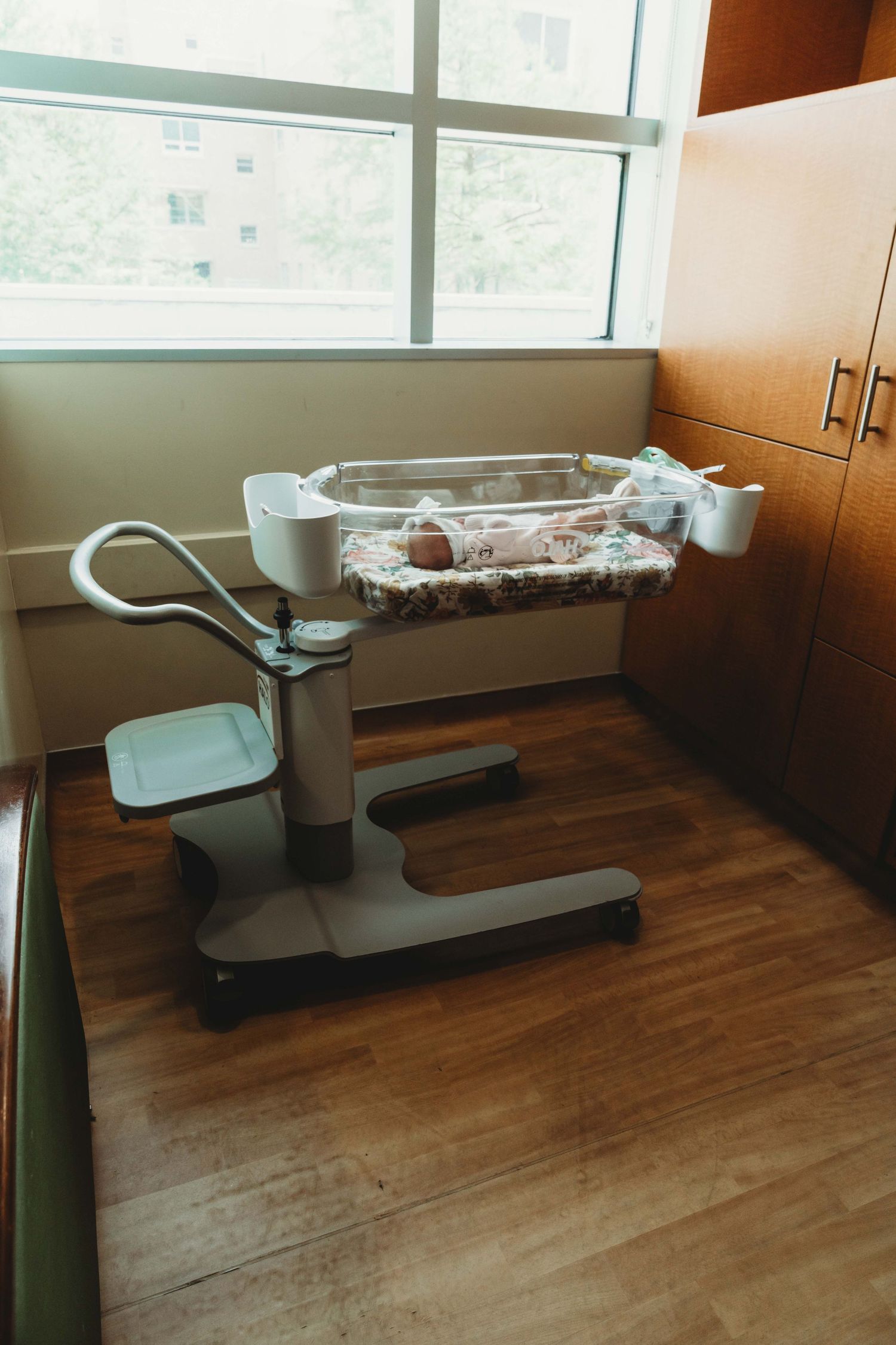A modern hospital bassinet containing a newborn rests near a window in a medical room with wooden floors.