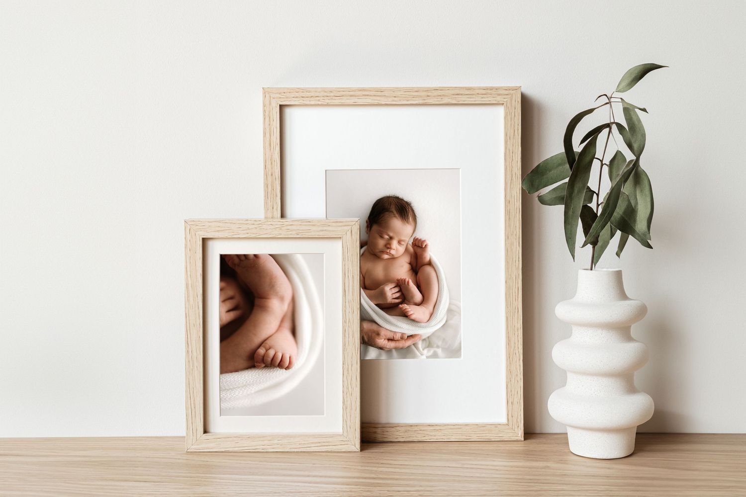 Framed newborn photos displayed with decorative vase on wooden surface.