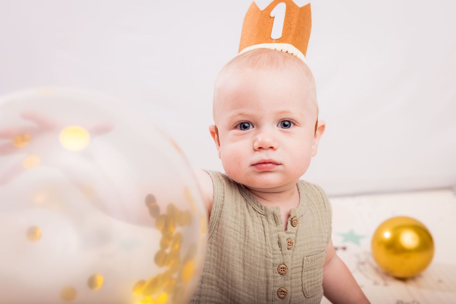 Baby wearing paper crown sits among gold and white party decorations.
