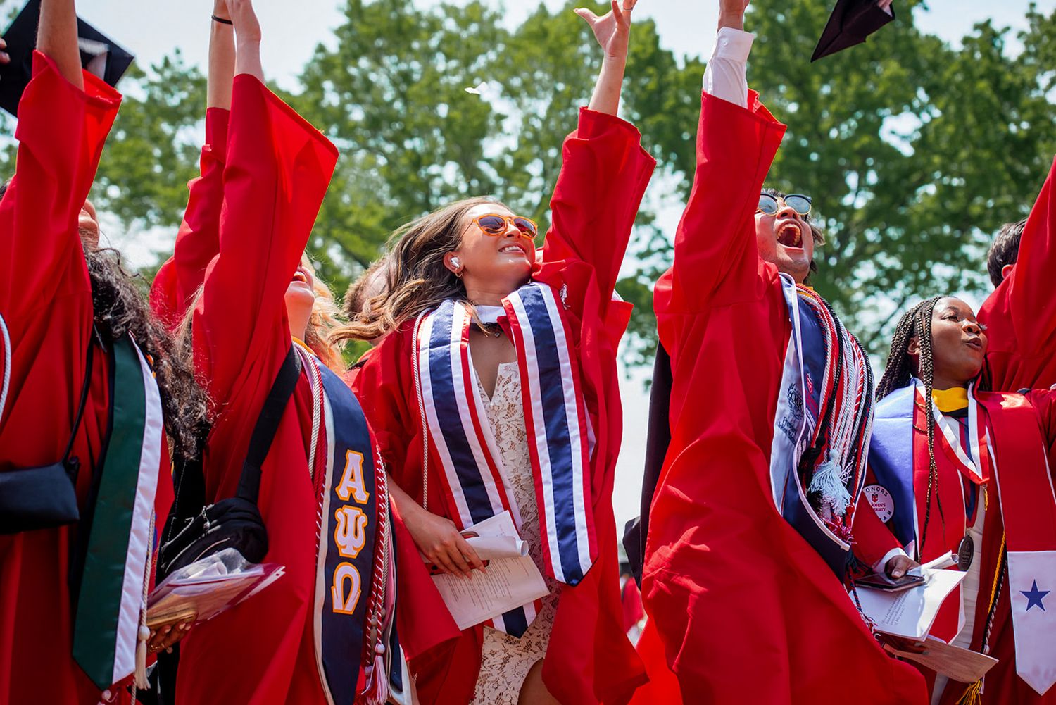 Graduates in bright red and striped regalia celebrate with raised arms during outdoor commencement ceremony.