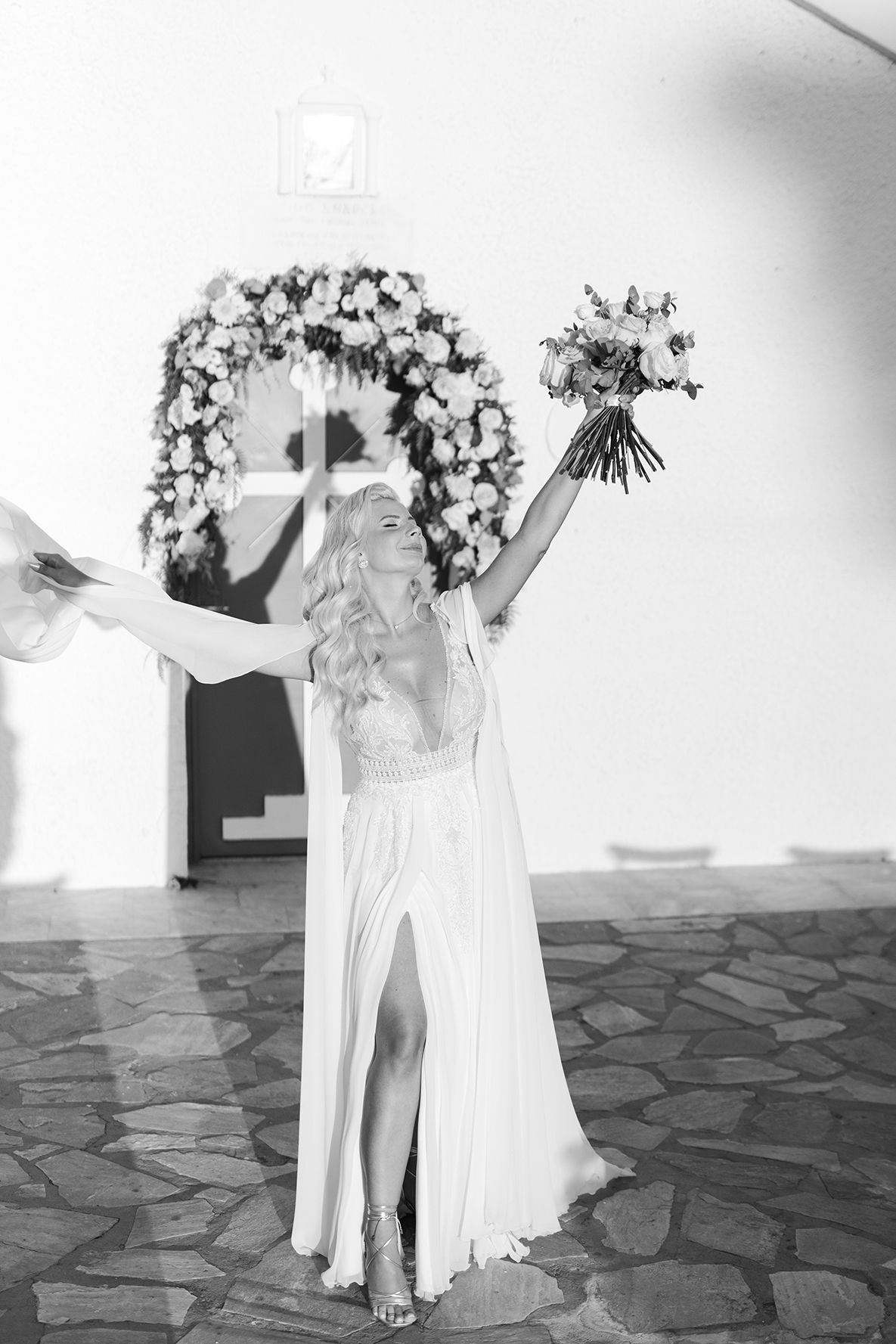 A bride in a flowing white dress with a high slit raises her bouquet joyfully under a floral arch in black and white. tonia and theodore