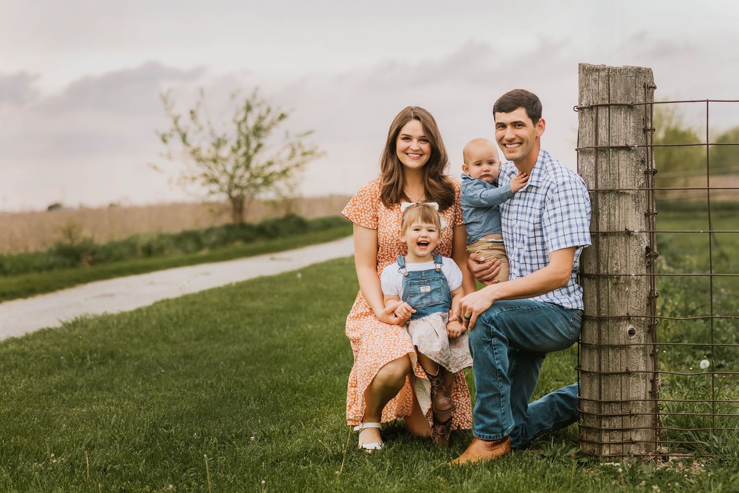 Family poses together on a sunny day in a grassy field with rustic fencing.
