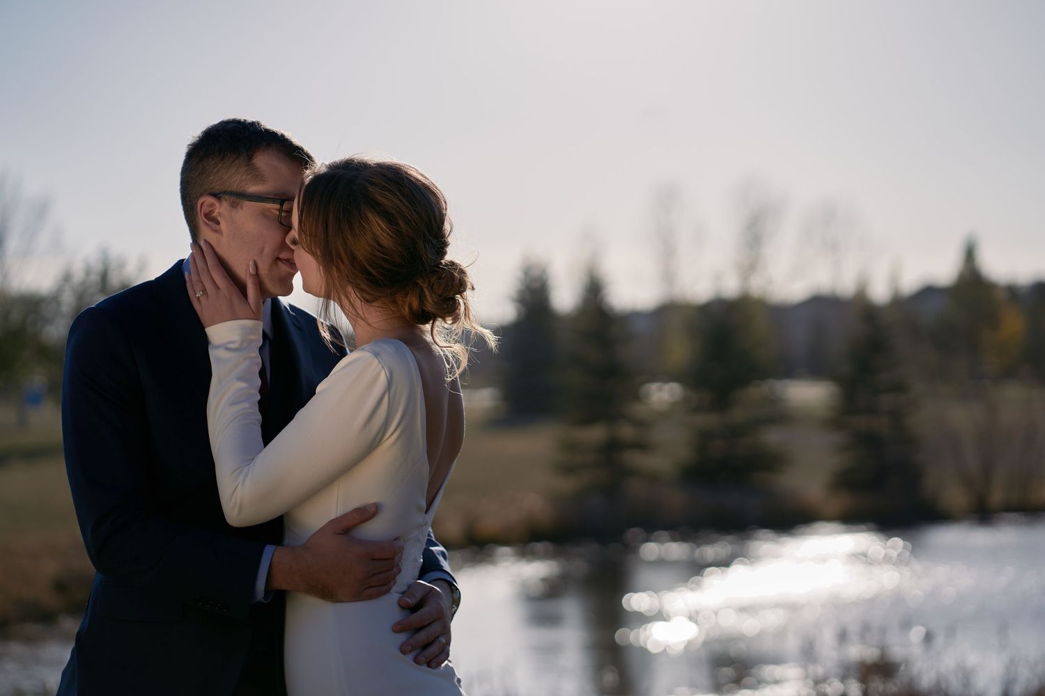 Wedding couple sharing an intimate moment by a lake at sunset with sunlight reflecting off the water's surface in Spruce Grove, Alberta.