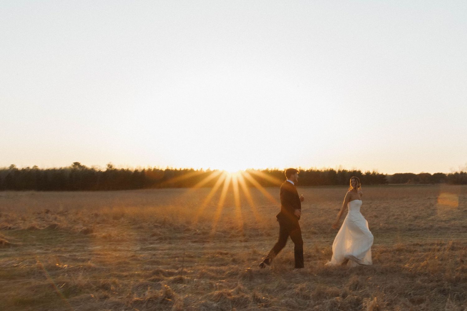 A dramatic sunset silhouettes a couple walking across an open field during their wedding photoshoot.