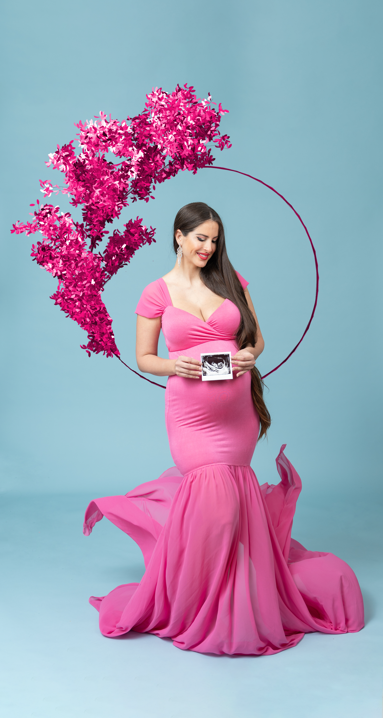 A woman in a flowing pink maternity dress poses elegantly with bright magenta flowers against a blue backdrop.