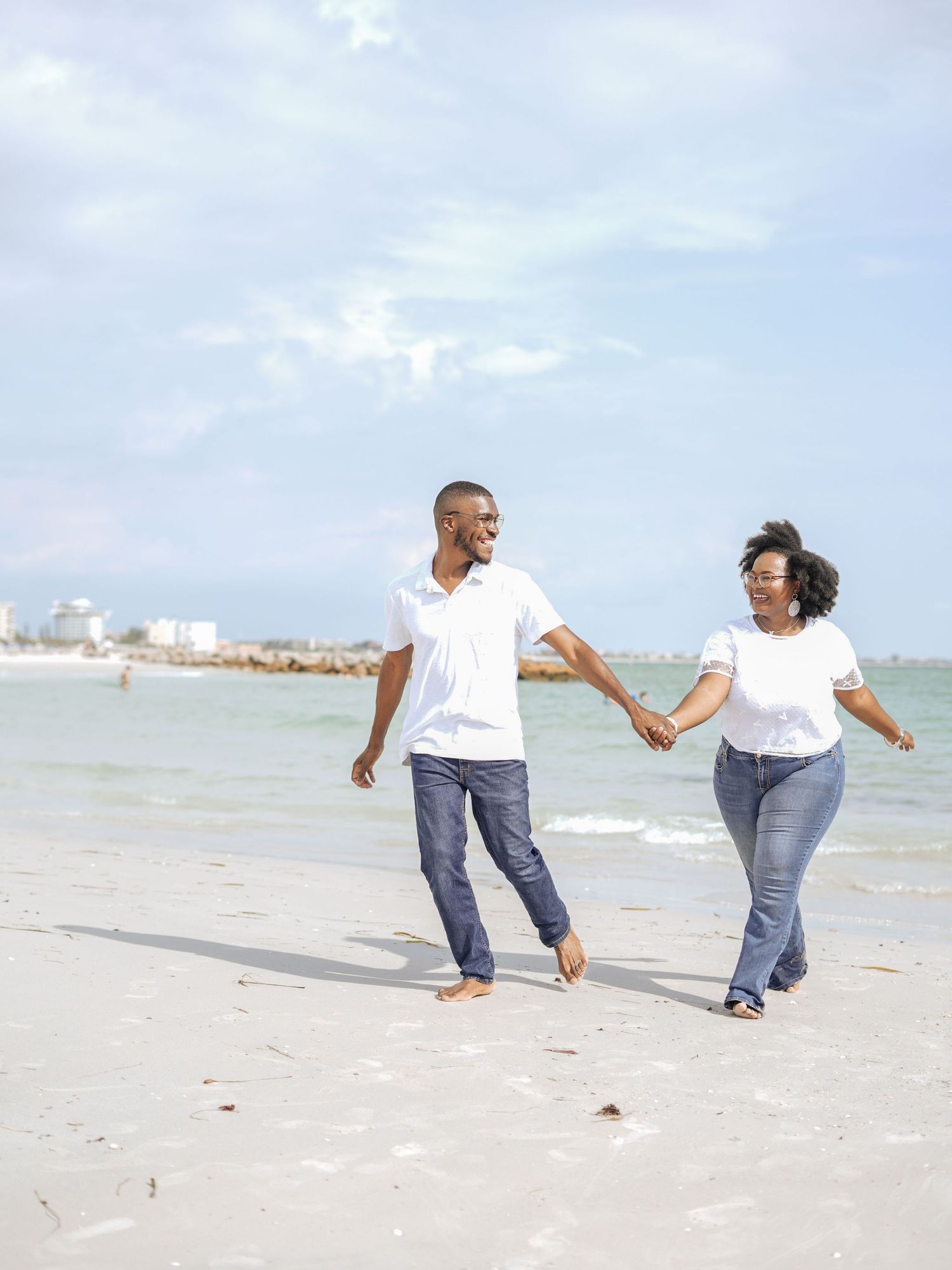 Couple holding hands while walking joyfully along a beach with cityscape in background.