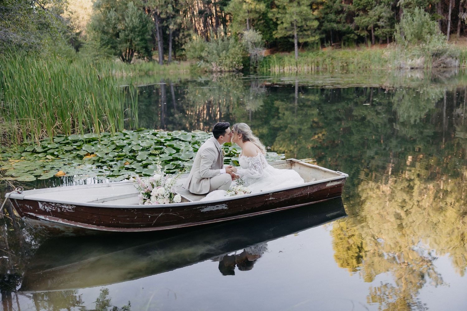 Romantic couple in formal attire share an intimate moment on a wooden dock surrounded by lily pads and reeds on a lake.