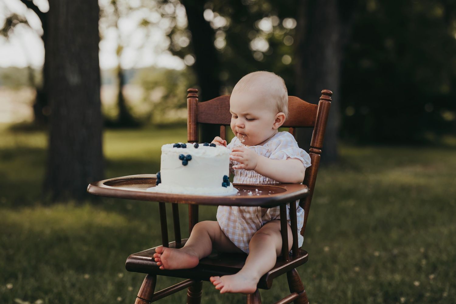 Young child in white dress sitting in wooden high chair during outdoor photoshoot.
