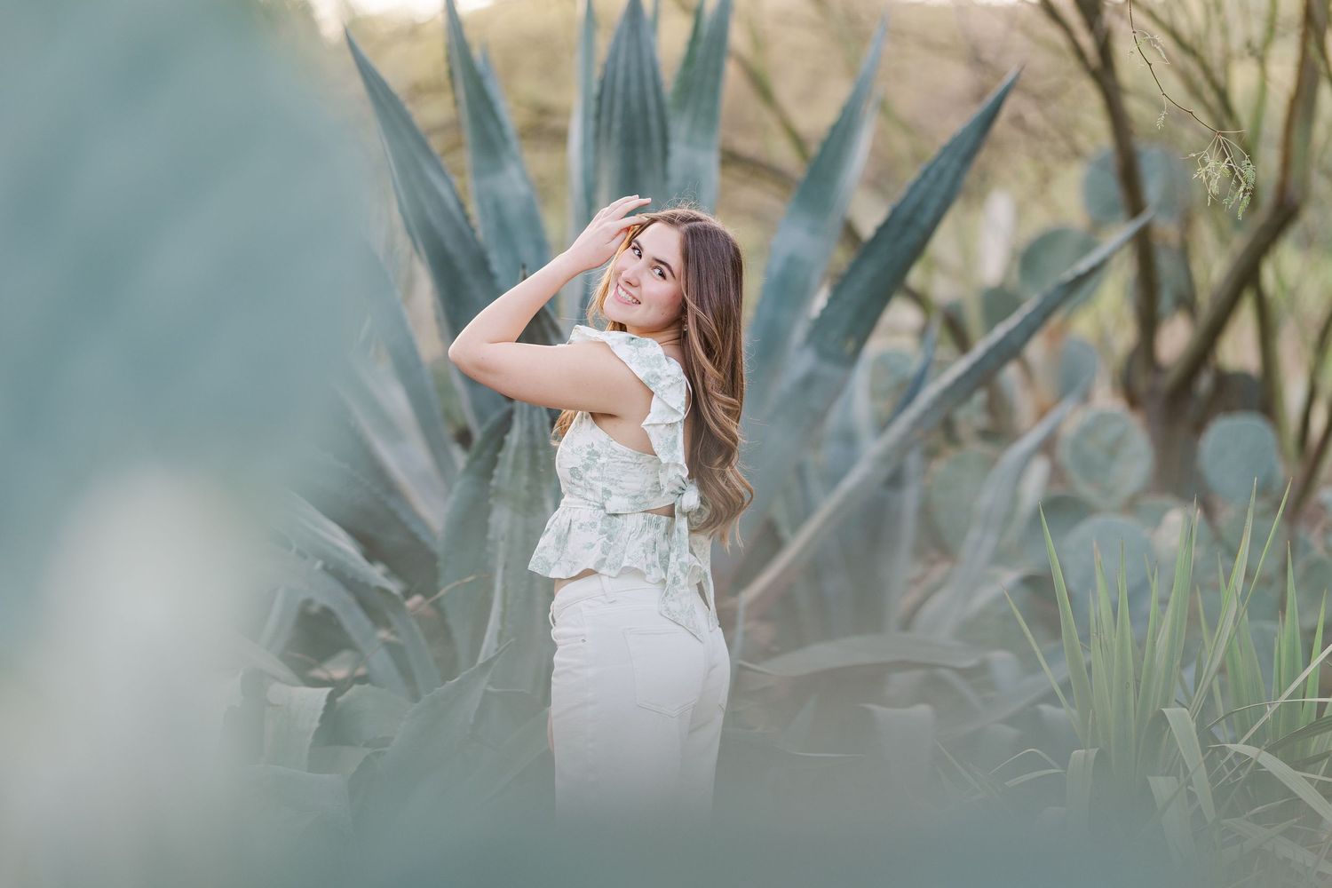 A person in a white dress poses elegantly among large agave plants in a dreamy outdoor setting.