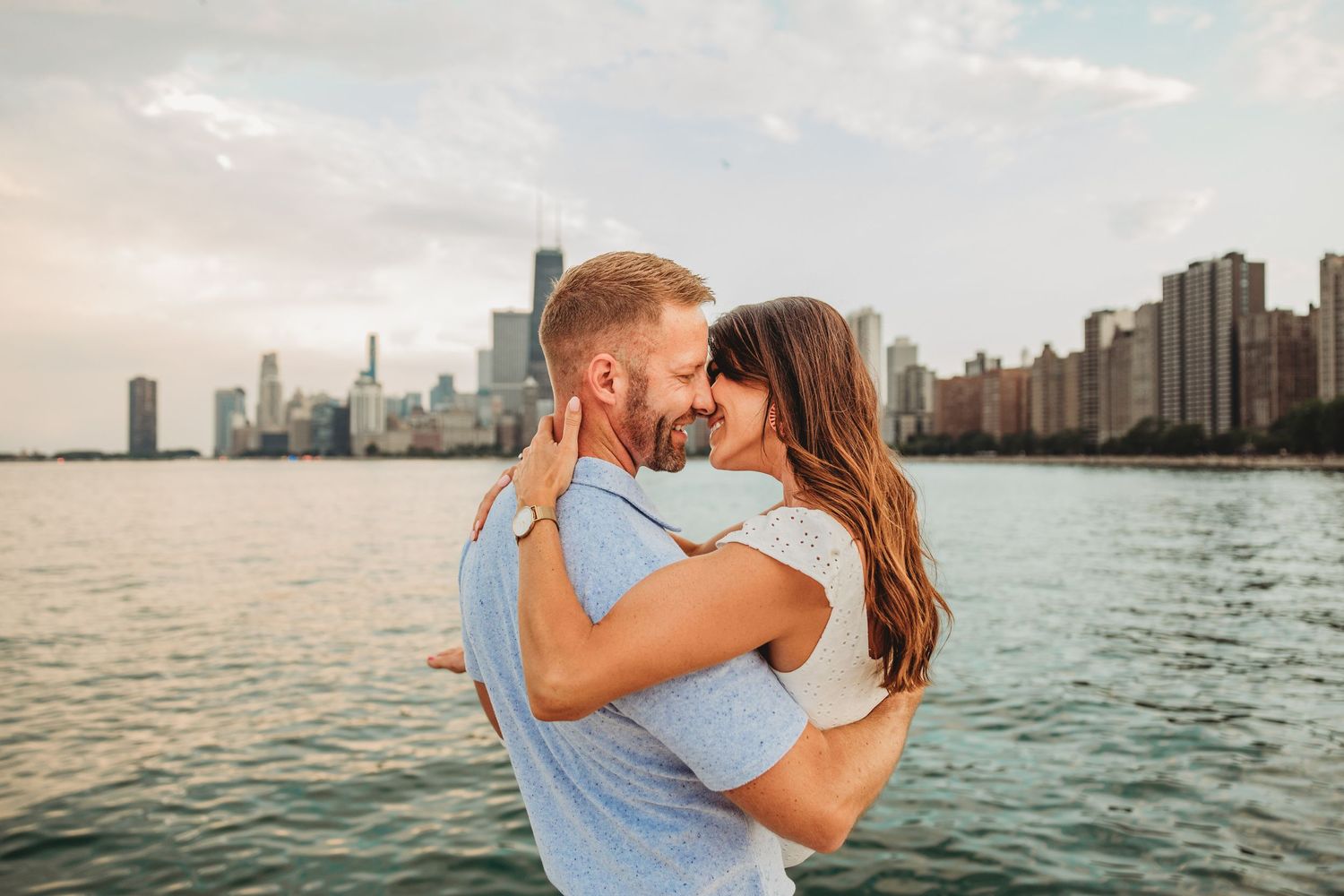 Loving couple poses together on beach with downtown Chicago skyline visible in distance.