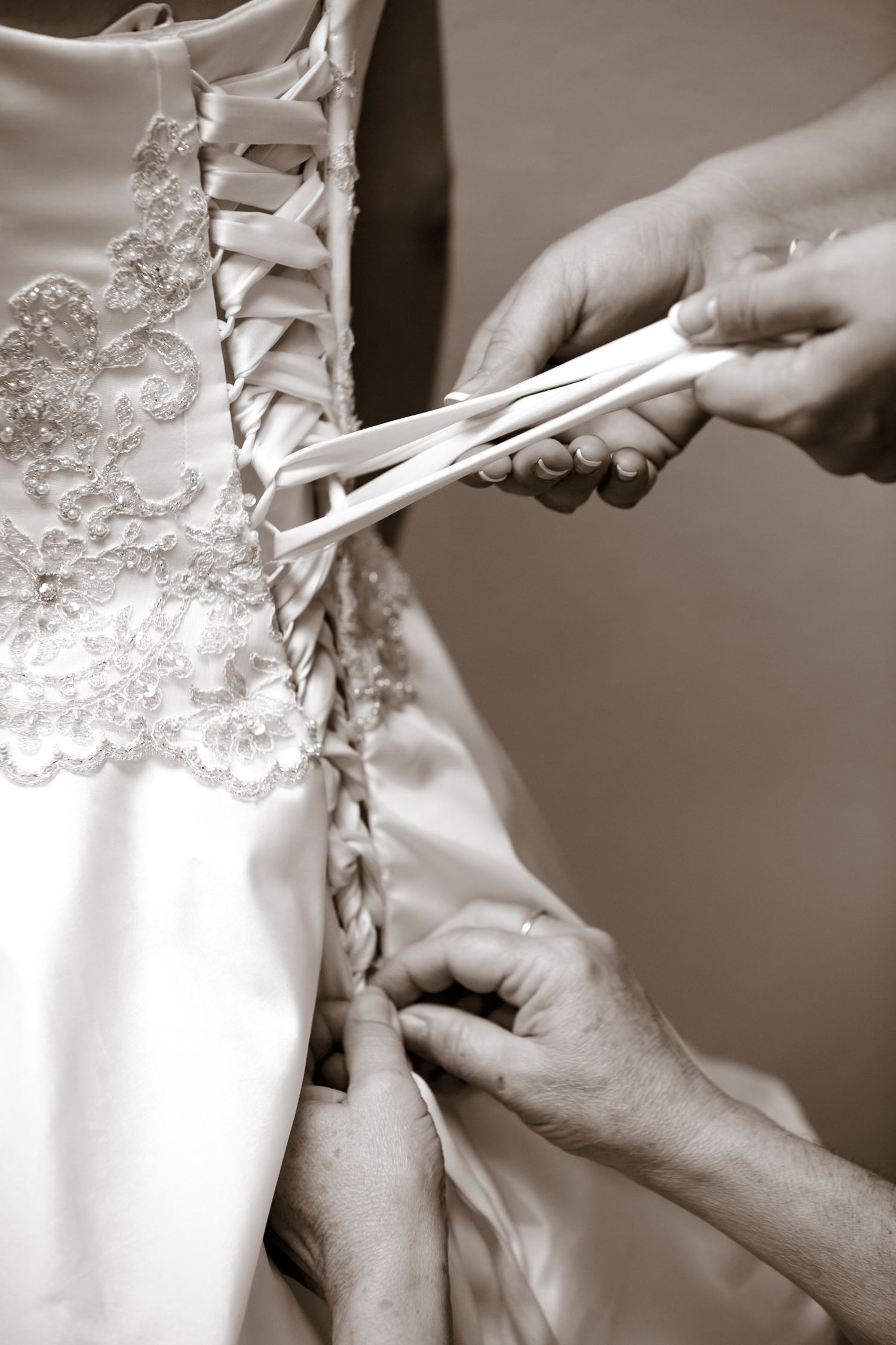 Hands lacing up the back of an ornate white wedding dress with satin ribbon and delicate lace details in sepia tone.