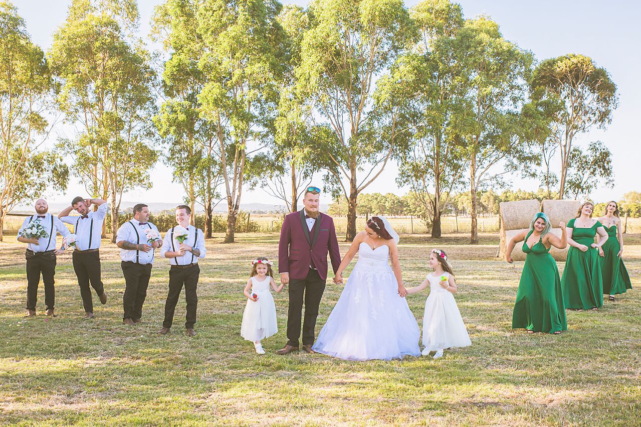 A wedding ceremony takes place outdoors in a grassy field with guests seated on white chairs under bright sunlight.