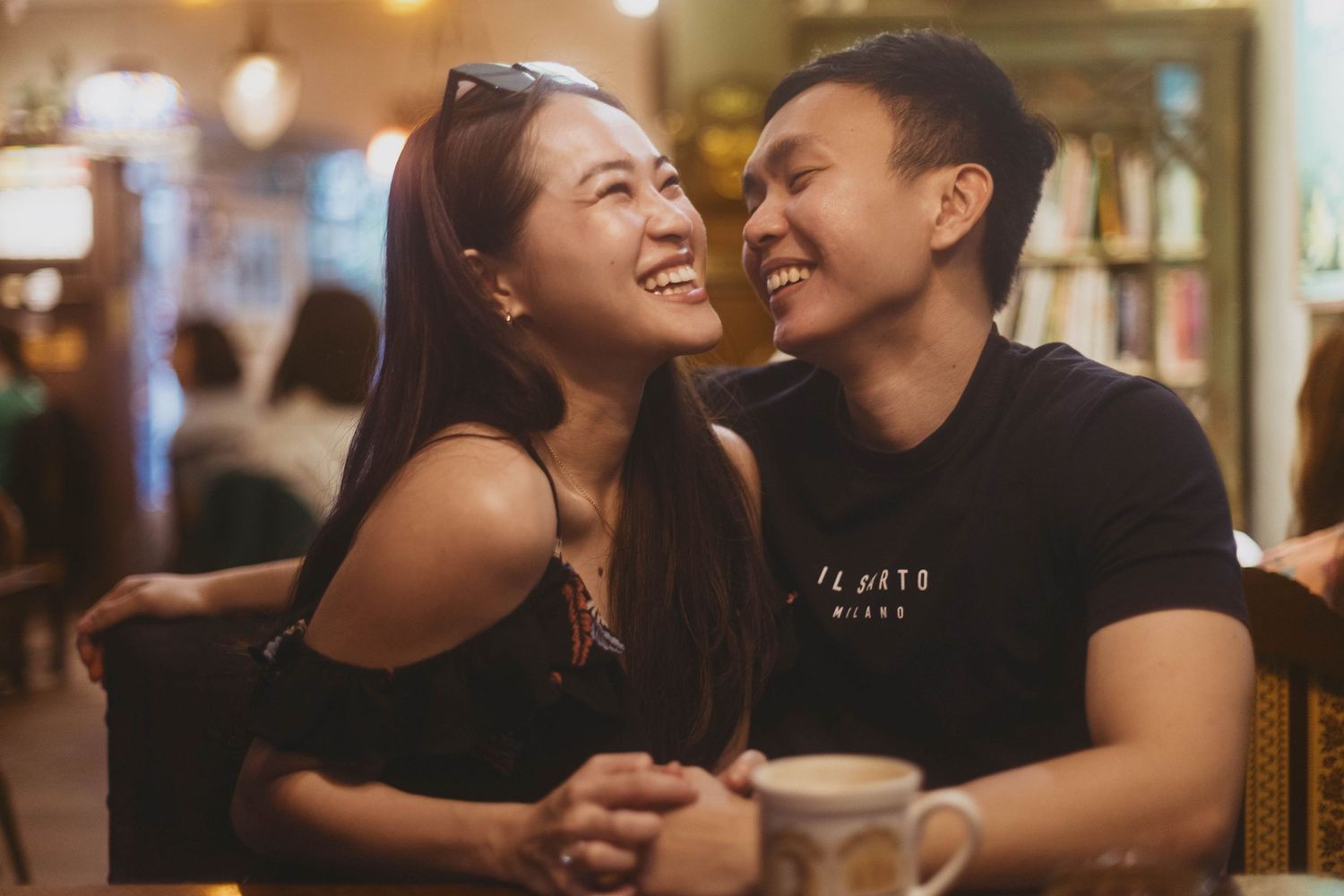 Happy Asian couple smiling and holding hands at a cozy cafe table in warm ambient lighting.