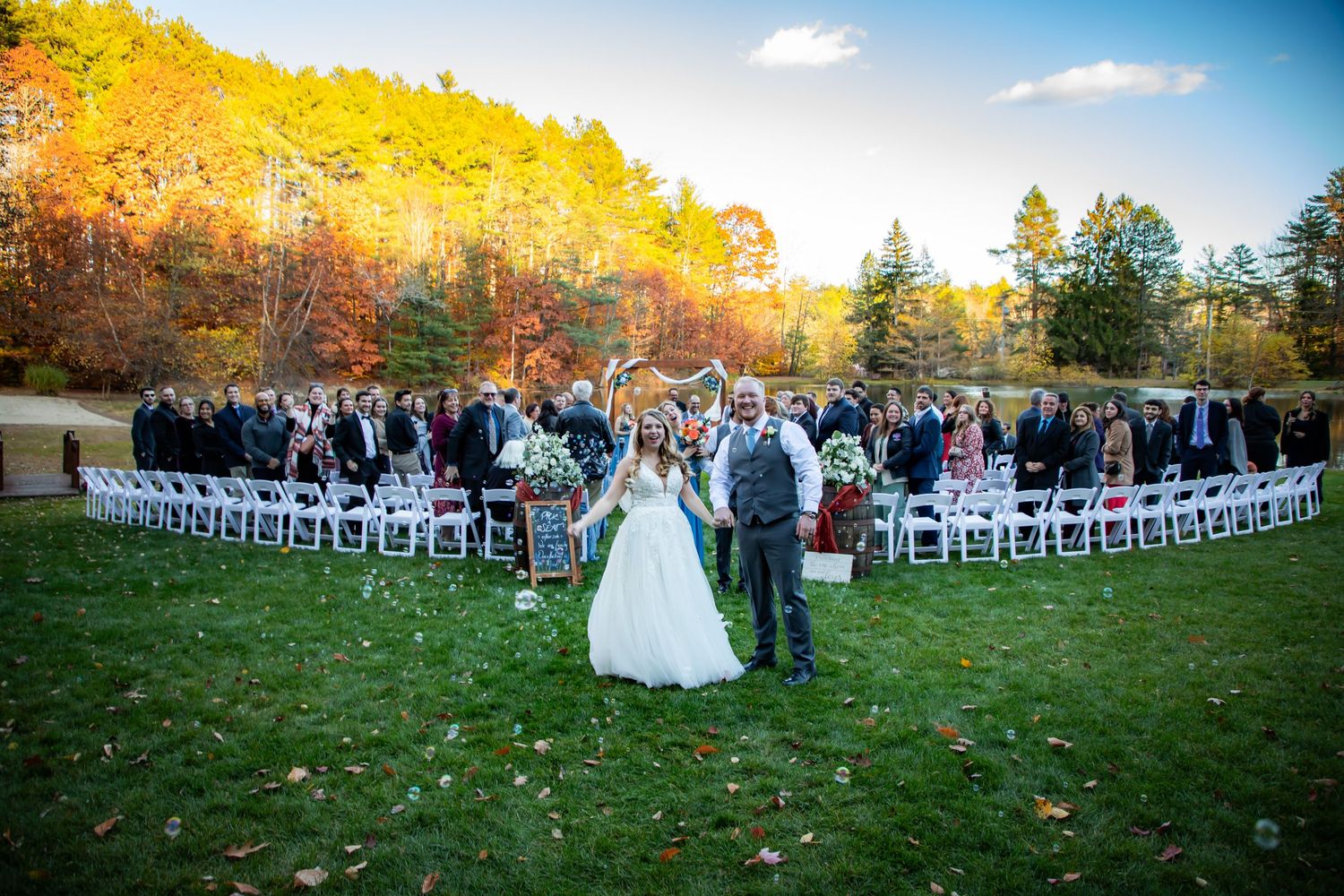 Outdoor fall wedding ceremony with white chairs and autumn foliage backdrop.