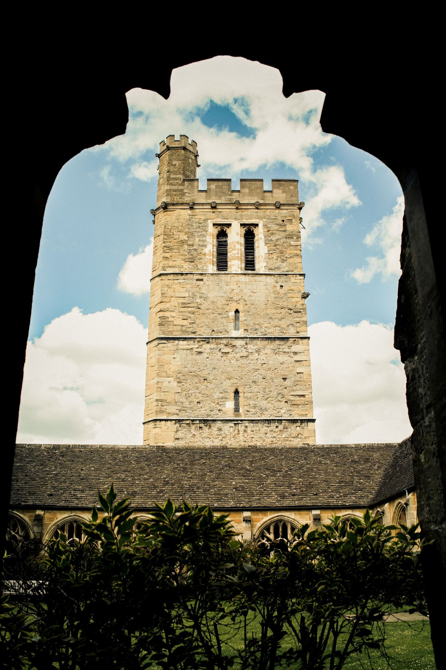 Ancient stone church tower with crenellations viewed through an archway against a cloudy blue sky in England.